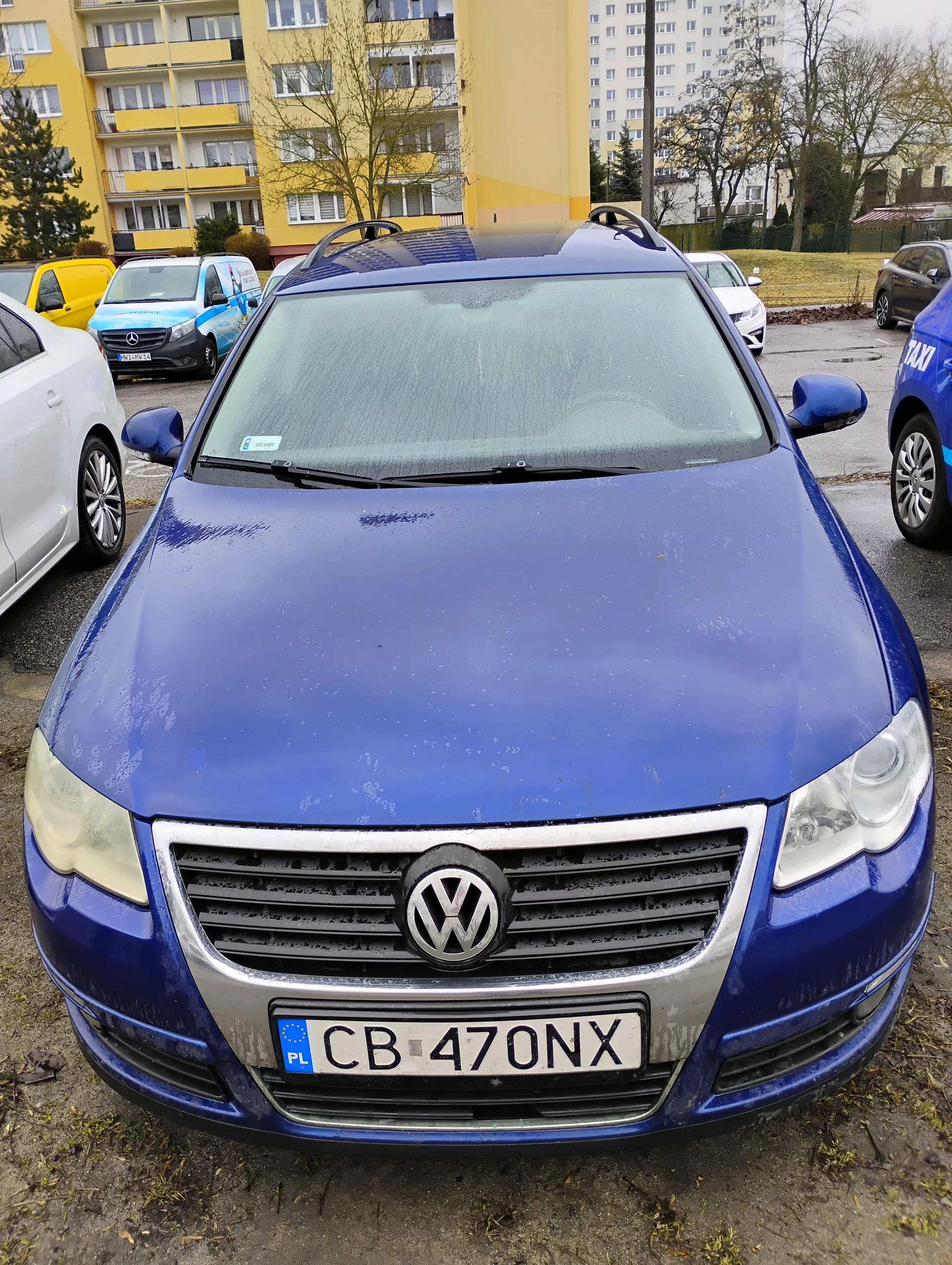 Niebieski samochód marki Volkswagen z tablicą rejestracyjną CB 470NX PL, zaparkowany na ulicy. W tle widoczne są inne pojazdy, w tym biały, żółty i niebieski samochód z napisem 'TAXI', budynki mieszkalne oraz drzewa bez liści. Na tablicy rejestracyjnej widoczny jest napis 'CB 470NX' oraz oznaczenie 'PL'.