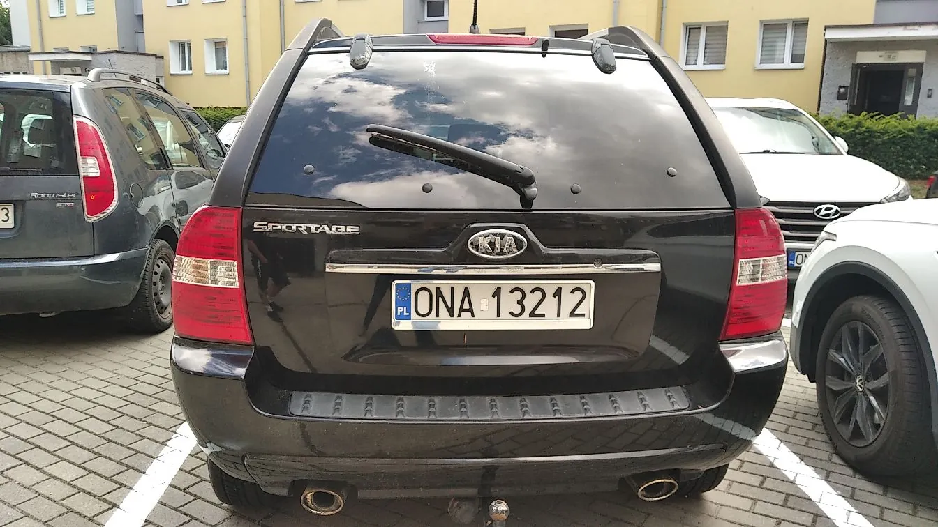 Tylny widok czarnego samochodu marki Kia Sportage zaparkowanego na kostce brukowej. Na klapie bagażnika widoczne jest logo KIA, napis SPORTAGE oraz tablica rejestracyjna o treści ONA 13212 z oznaczeniem PL. Po lewej stronie stoi ciemnoszary samochód, a po prawej biały samochód. W tle widoczny jest żółty budynek mieszkalny.