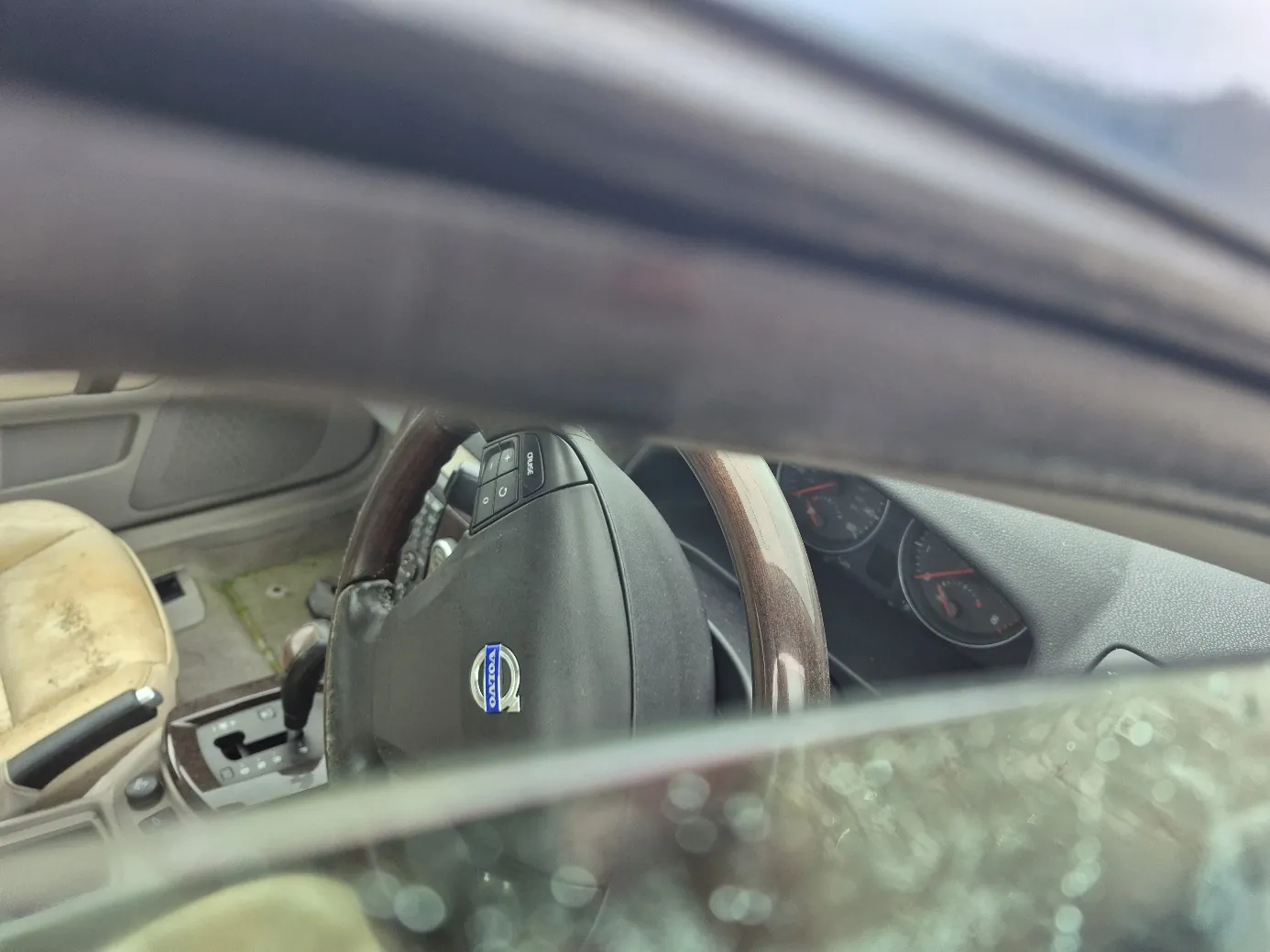 Wnętrze samochodu Volvo widziane przez przednią szybę. Widoczna kierownica z logo Volvo, deska rozdzielcza z dwoma okrągłymi wskaźnikami, beżowe siedzenia z widocznymi zabrudzeniami, dźwignia zmiany biegów oraz fragment przedniej szyby z plamami. W tle rozmyty krajobraz z górami.