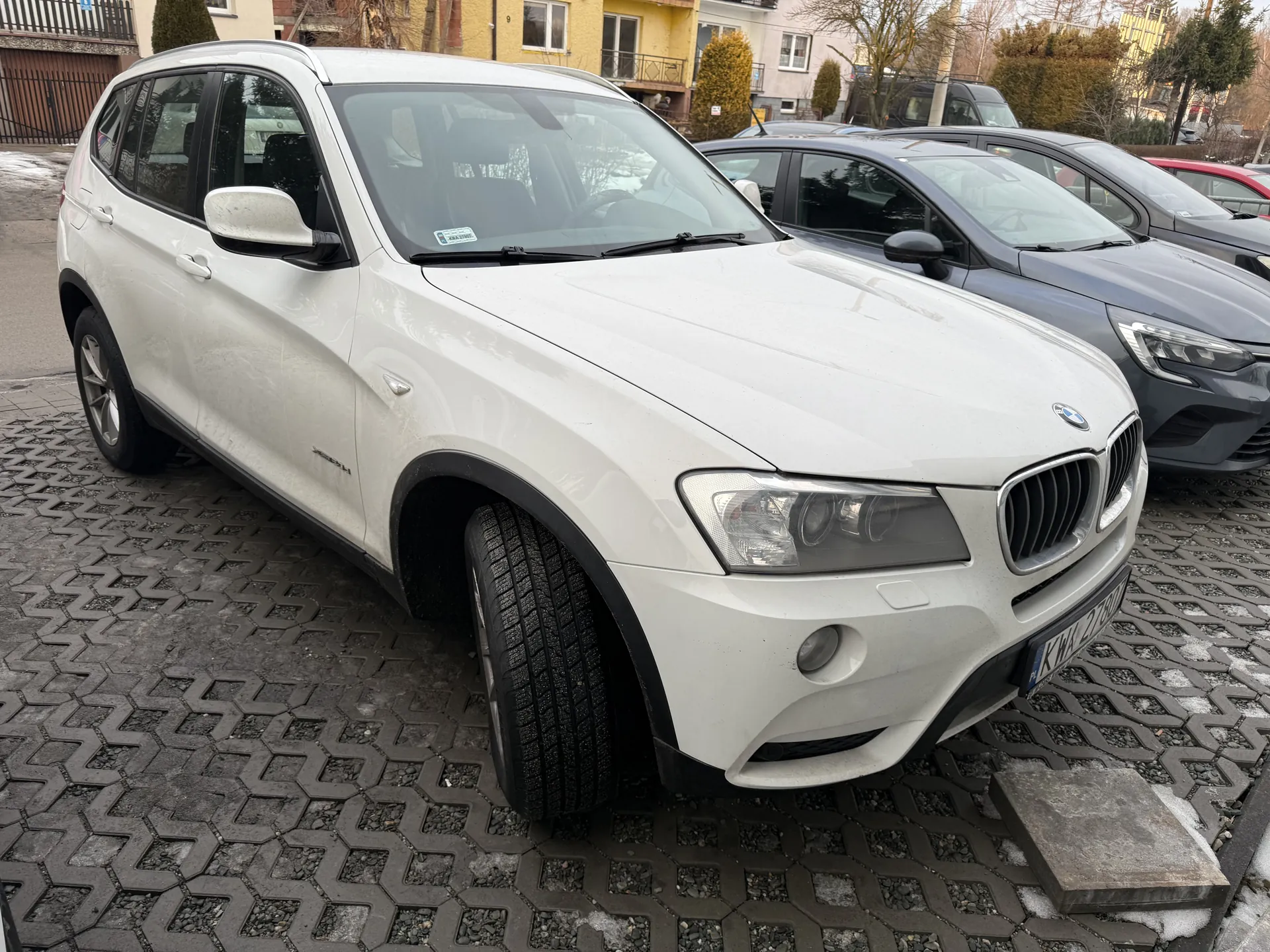 Biały samochód SUV marki BMW X3 z tablicą rejestracyjną KTW 2780 zaparkowany na podwórku z kostkowanym asfaltem. W tle widoczne są inne pojazdy (szary sedan i czerwony samochód), budynki mieszkalne z żółtymi i białymi elewacjami, krzewy oraz resztki śniegu na ziemi. Na przedniej szybie BMW znajduje się naklejka. Samochód ma czarne obwiednie nadkoli i opony zimowe.