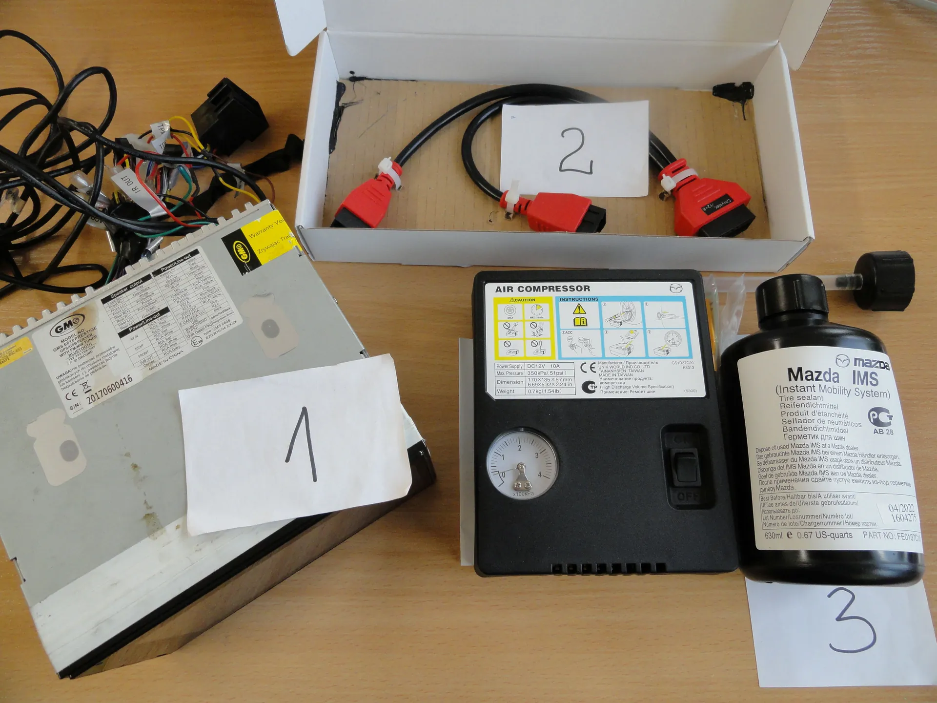 Na drewnianym stole umieszczono różne elementy elektroniczne. Po lewej stronie srebrny aparat z etykietą 'GM', numerem seryjnym '20170600416' i żółtym naklejką, przykryty białym kartonikiem z cyfrą '1'. Obok niego splątane czarne i kolorowe przewody z złączami. W centrum otwarte białe pudełko z czerwonymi złączami, czarnymi kablami i kartonikiem z cyfrą '2'. Przy pudełku czarny aparat oznaczony 'AIR COMPRESSOR' z manometrem (skala 0-4), instrukcjami i przełącznikiem 'ON/OFF'. Po prawej czarna butelka z etykietą 'Mazda IMS (Instant Mobility System)', informacją '630ml e 0.67 US-quarts' oraz kartonikiem z cyfrą '3'.