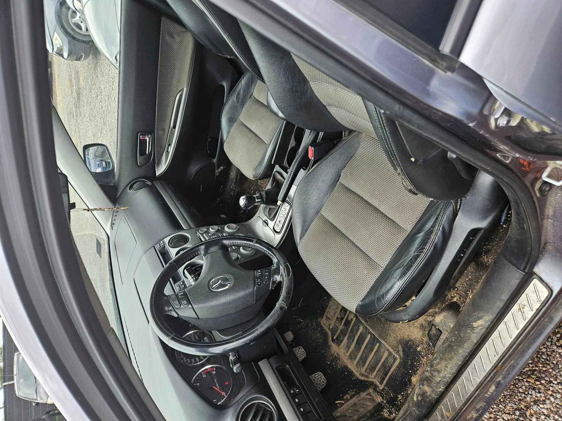 Wnętrze samochodu widziane z perspektywy otwartych drzwi kierowcy. Widoczna jest czarna kierownica z logo Mercedes-Benz, deska rozdzielcza, dwa przednie fotele z jasnoszarą materiałową tapicerką w centralnej części i czarnymi skórzanymi bokami. Podłoga samochodu jest zabrudzona piaskiem i ziemią.