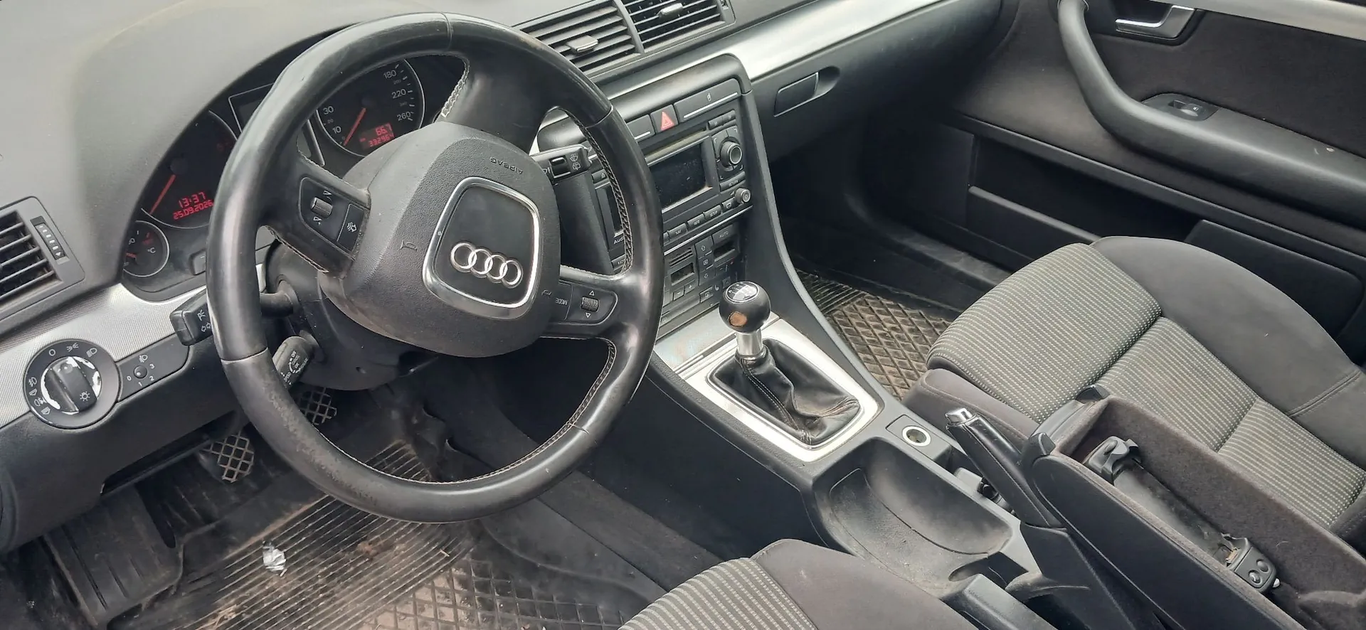 Wnętrze samochodu marki Audi z perspektywy kierowcy. Widoczna jest czarna skórzana kierownica z logo Audi w centralnej części, deska rozdzielcza z radiem i panelami sterowania, wskaźniki prędkości oraz obrotomierza z czerwonymi cyframi. W konsoli środkowej znajduje się drążek zmiany biegów manualnej. Widoczne są dwa przednie fotele z szarym, prążkowanym materiałem oraz czarne gumowe dywaniki na podłodze.