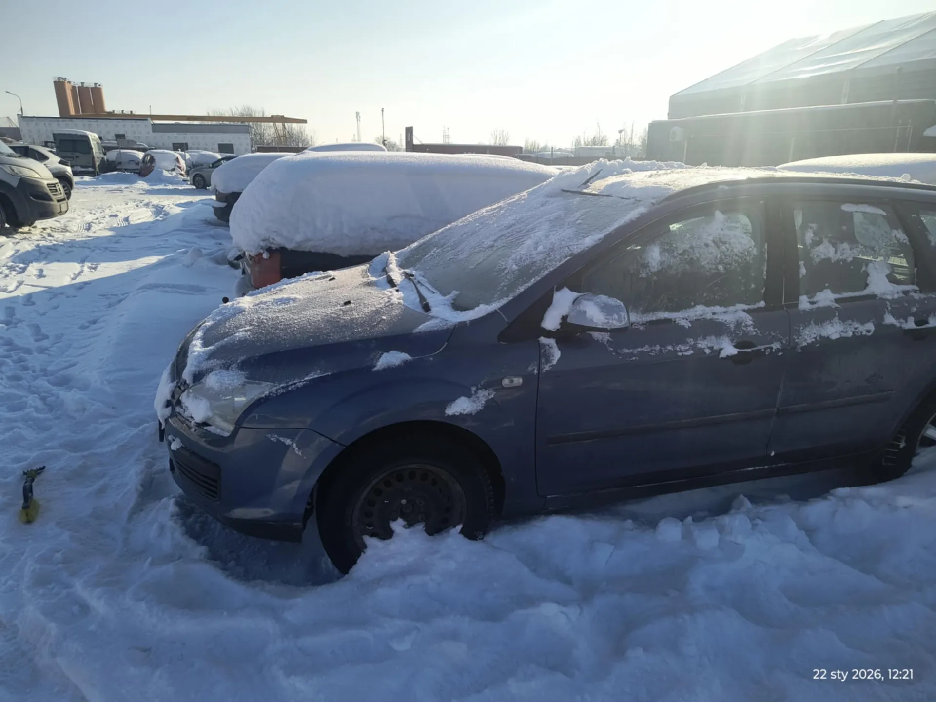 Zdjęcie przedstawia parking pokryty śniegiem z kilkoma samochodami. Na pierwszym planie niebieski samochód osobowy zasłany śniegiem, widoczne są również inne pojazdy w tle, częściowo zakryte śniegiem. Tło stanowią budynki przemysłowe, słoneczna pogoda i jasne niebo. W prawym dolnym rogu widoczny znaczek z datą i czasem: 22 sty 2026, 12:21.
