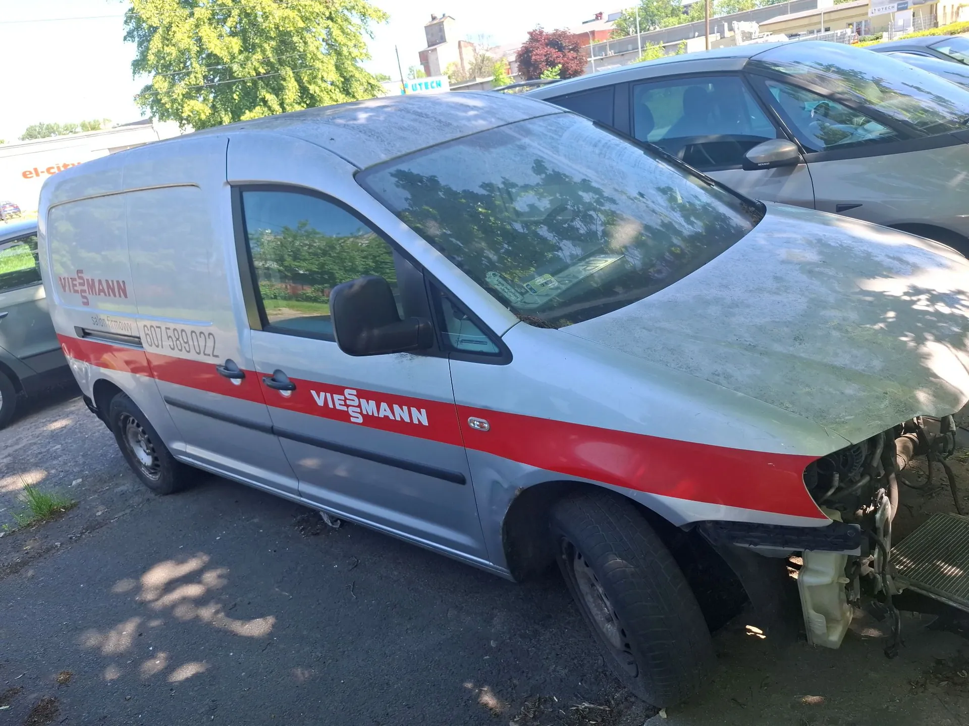 Srebrny samochód dostawczy marki Volkswagen z czerwonym pasem na boku i napisem 'VIEßMANN' oraz numerem telefonu '607 589 022'. Pojazd jest uszkodzony w przedniej części po prawej stronie, brakuje zderzaka i maska jest wgnieciona. Samochód stoi zaparkowany na asfaltowym podjeździe w pobliżu zielonych drzew.