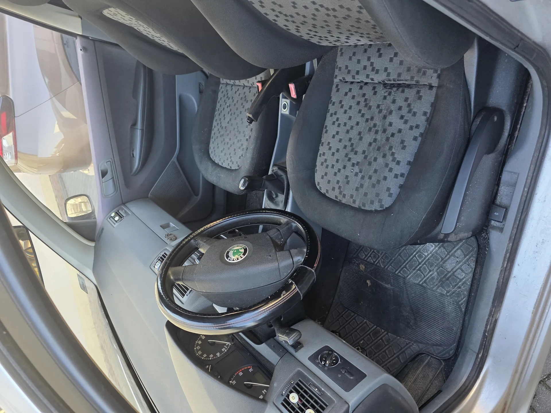 Wnętrze samochodu z widocznym kierownicą z logo Škoda, czarnymi siedzeniami w szachownicowy wzór, deską rozdzielczą z licznikami, czarnymi dywanikami podłogowymi oraz otwartą drzwiami. W tle widoczna część srebrnego nadwozia i odbicie innego pojazdu w lusterku bocznym.