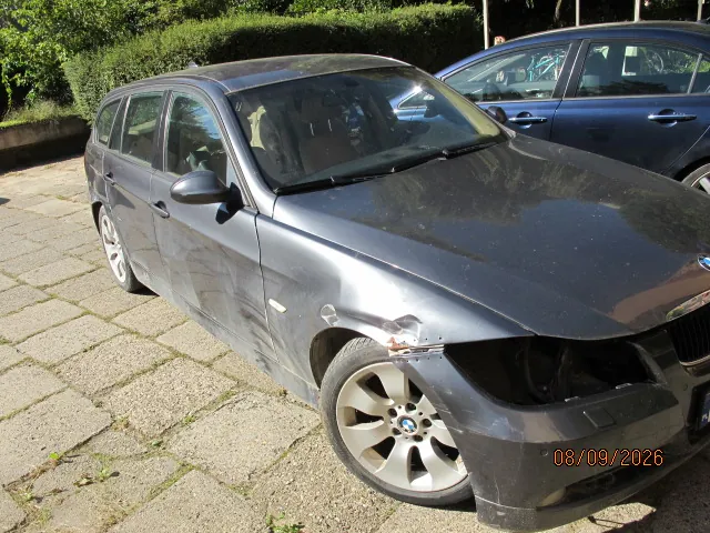Czarny samochód marki BMW w nadwoziu kombi, zaparkowany na kostce brukowej. Pojazd jest uszkodzony: widoczne są wgniecenia na przednim prawym błotniku oraz na prawych drzwiach. Brakuje prawego reflektora przedniego. W tle widoczny jest fragment innego, ciemnego samochodu oraz zielone krzewy. W prawym dolnym rogu zdjęcia znajduje się pomarańczowy napis z datą: 08/09/2026.