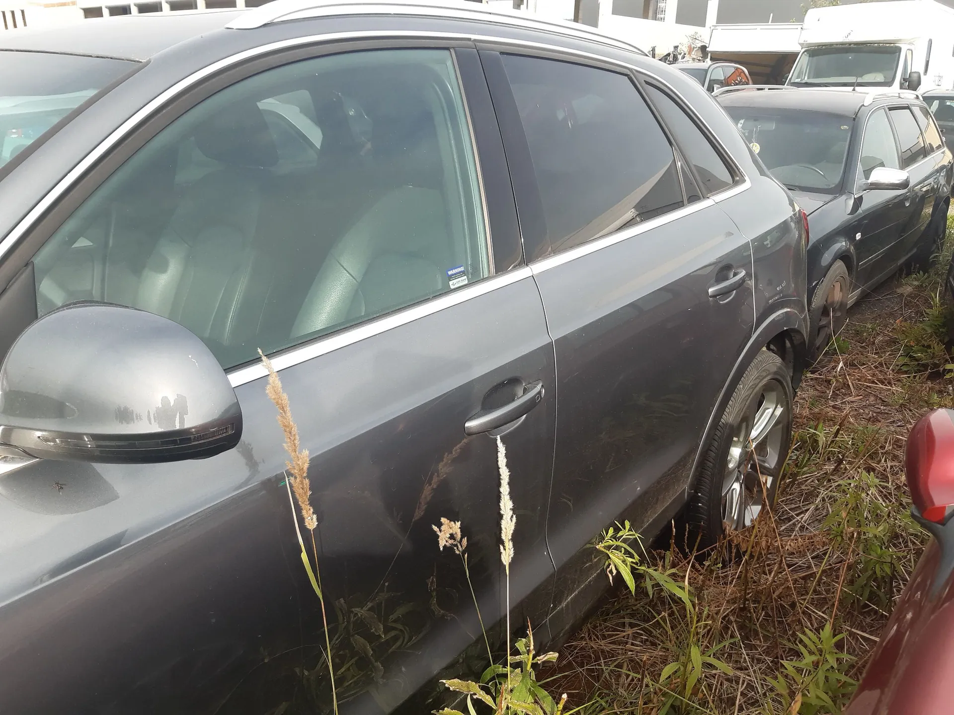 Zbliżenie na bok szarego samochodu typu SUV zaparkowanego na trawiastym terenie. Widoczne są drzwi kierowcy, lusterko boczne oraz przednie koło z aluminianą felgą. W tle widać inne zaparkowane samochody, w tym czarny samochód osobowy i biały pojazd dostawczy.