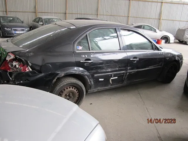 Czarny samochód osobowy typu sedan z widocznymi uszkodzeniami w tylnej lewej części: zniszczony tylny zderzak, uszkodzony reflektor i wgniecenia nad kołem. Na tylnej szybie naklejka w kształcie flagi Stanów Zjednoczonych. Samochód stoi w garażu lub hali z metalowymi ścianami, w tle widoczne są inne pojazdy, w tym srebrny Audi i biały samochód. W prawym dolnym rogu obrazu widoczny jest napis 14/04/2026. Podłoga betonowa, oświetlenie naturalne.