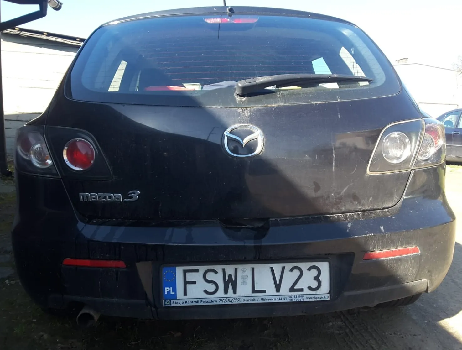 Tył czarnego samochodu Mazda 3 z widocznym logo marki, napisem "mazda 3" po lewej stronie bagażnika oraz tablicą rejestracyjną o numerze FSW LV23 z kodem kraju PL. Na tablicy widoczny jest również napis "Stacja Kontroli Pojazdów MERCX" oraz adres. Samochód znajduje się na zewnątrz, na tle budynku i częściowo widocznego innego pojazdu. Bagażnik i tylna szyba mają ślady zabrudzeń.