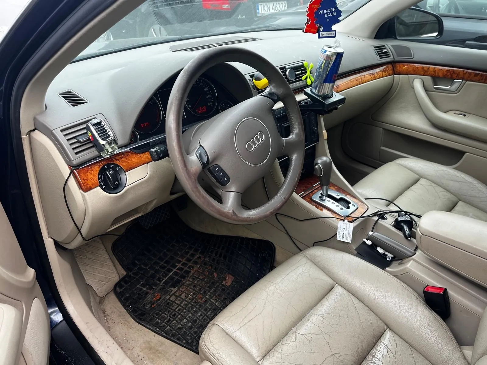 Wnętrze samochodu marki Audi z kierownicą z logo czterech pierścieni. Deska rozdzielcza w kolorze beżowym z brązowymi wstawkami drewnianymi. Na desce rozdzielczej widoczny napój Red Bull w uchwycie, żółty i zielony akcesorium oraz powiewnik w kształcie drzewka z napisem "WUNDER-BAUM". Przez przednią szybę widoczna tablica rejestracyjna TKN 46328. Siedzenia tapicerowane beżową skórą z widocznymi pęknięciami. Czarne dywaniki podłogowe z brudnymi plamami. W otwartym drzwiach widoczny uchwyt i panel boczny. Przełącznik biegów umieszczony na środkowej konsoli.