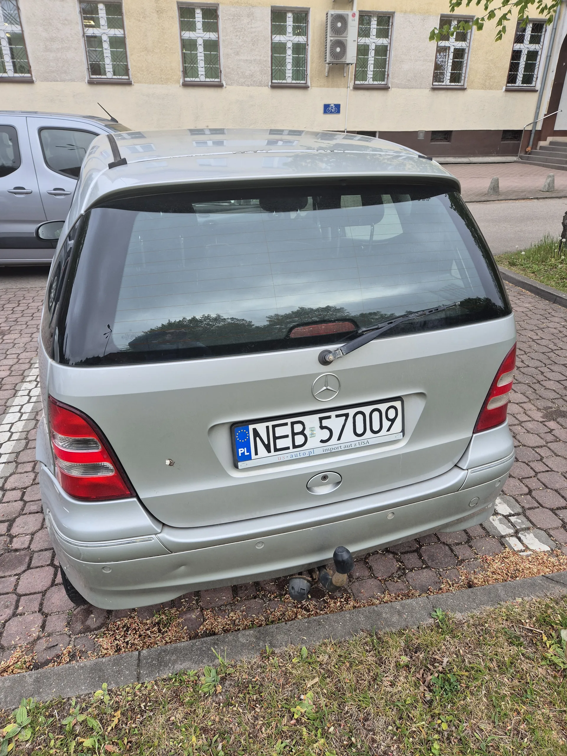Srebrny samochód Mercedes-Benz widziany od tyłu z tablicą rejestracyjną NEB 57009 PL. Samochód zaparkowany na brukowanej ulicy. Po lewej stronie budynki z oknami i klimatyzatorami, po prawej trawnik z suchymi liśćmi.