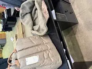 Wnętrze sklepu z widocznym beżowym pikowanym płaszczem z białą etykietą na czarnym stole. Obok płaszcza leży szara tkanina lub koc. W tle znajdują się półki z różnymi przedmiotami oraz betonowa podłoga. Brak czytelnego tekstu poza nieczytelną etykietą na płaszczu.
