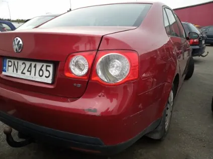 Czerwony samochód marki Volkswagen z widocznym tyłem, tablicą rejestracyjną PN-24165 oraz lekkimi uszkodzeniami na tylnym zderzaku. W tle inne zaparkowane pojazdy i czerwona budowla. Samochód stoi na asfaltowej powierzchni.