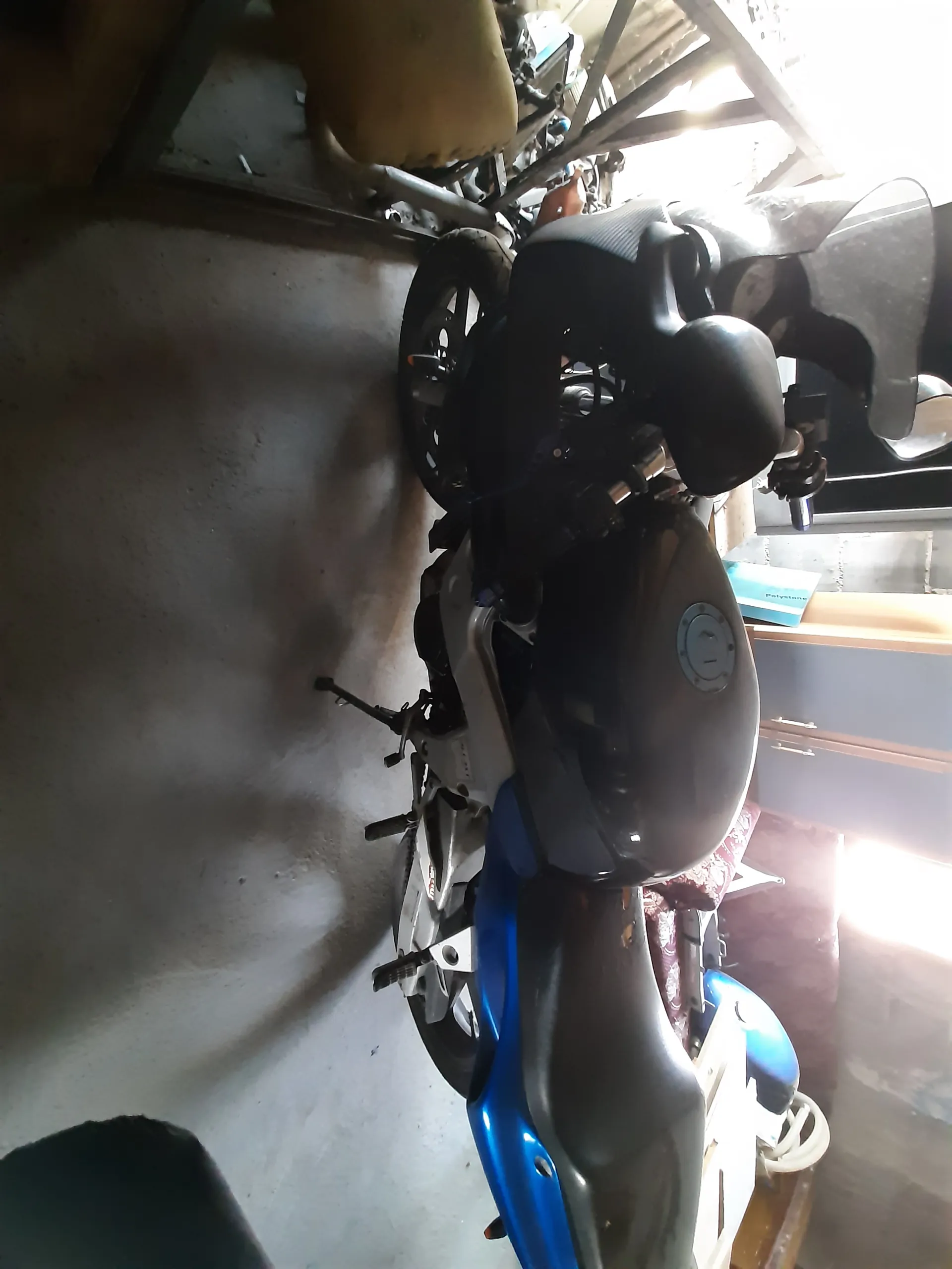 Czarno-niebieski motocykl zaparkowany w pomieszczeniu na betonowej podłodze. Motocykl stoi na podstawce. W tle widoczne są różne przedmioty, w tym czerwona sofa z wzorzystym pokrowcem, niebieska książka lub folder oraz narzędzia. Po lewej stronie znajduje się kask motocyklowy. Oświetlenie pochodzi z okien po lewej stronie obrazu.