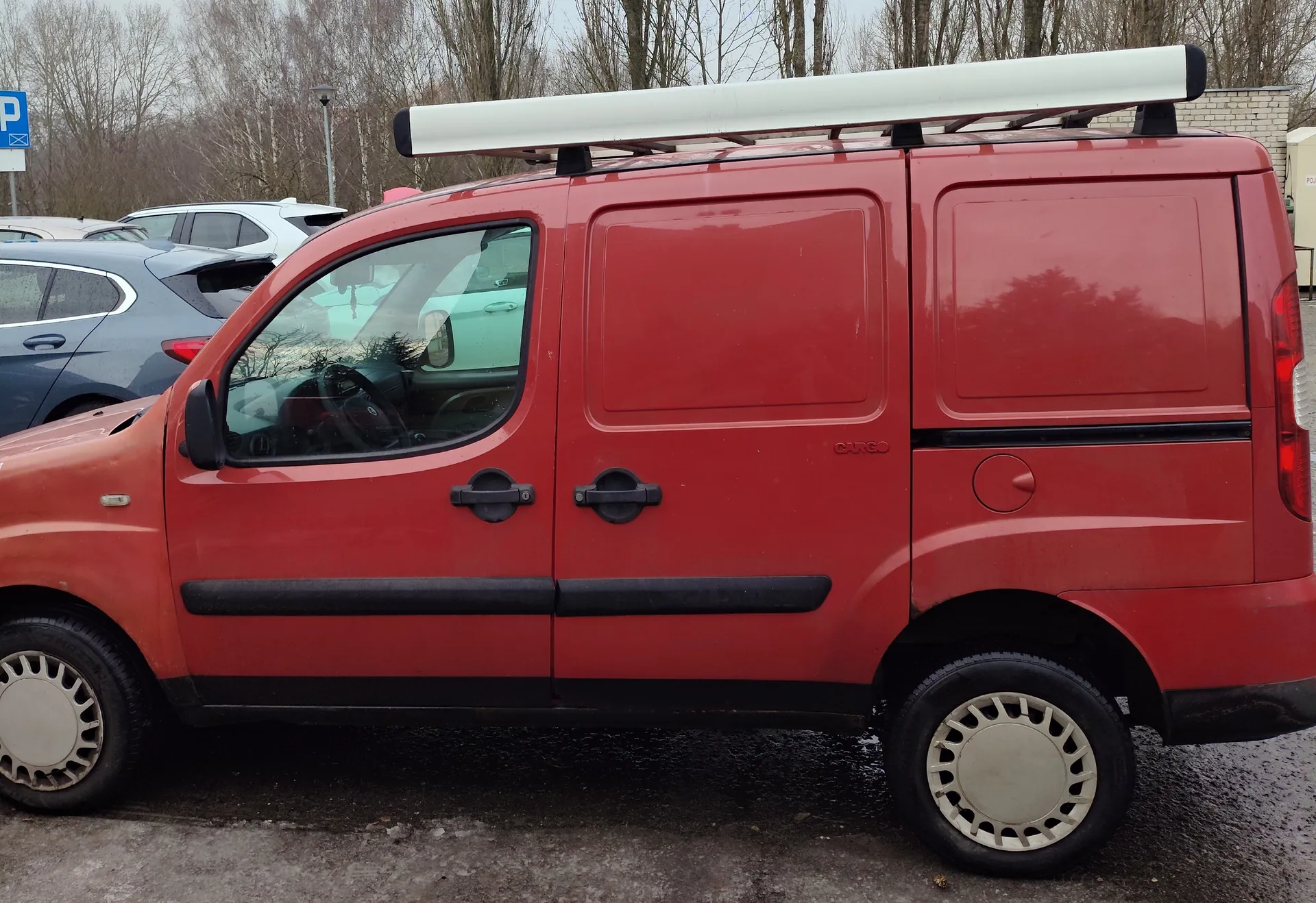 Czerwony samochód dostawczy z białym dachowym bagażnikiem, widok z boku. Na boku pojazdu widoczny napis "CARGO". Samochód ma czarne uchwyty drzwiowe i czarną listwę boczną. W tle zaparkowane są inne pojazdy, w tym niebieski samochód osobowy i biały SUV. Po lewej stronie widoczny niebieski znak parkingu z białą literą "P". Tło stanowią bezlistne drzewa i betonowa nawierzchnia. Podłoga wydaje się mokra.