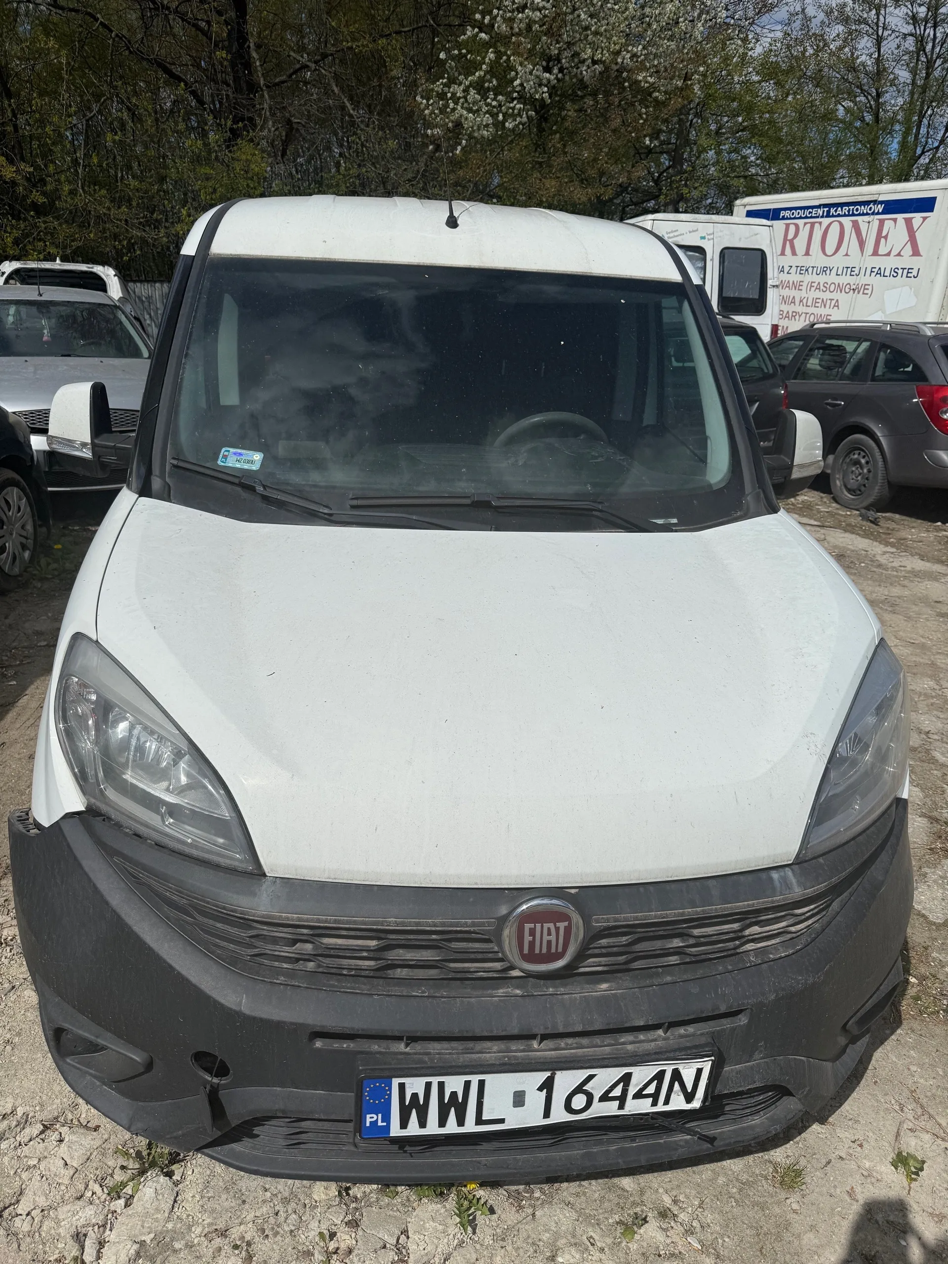 Biały samochód dostawczy marki Fiat z przodu, z widoczną tablicą rejestracyjną WWL 1644N z kodem PL. Na masce logo FIAT, przedni zderzak w kolorze czarnym. W tle inne pojazdy: srebrny samochód po lewej, ciemnoszary SUV po prawej oraz biały naczepowy pojazd z napisem RTONEX i dodatkowym tekstem w języku polskim. Tło stanowią drzewa i niebo. Podłoga z kamienistego gruntu.