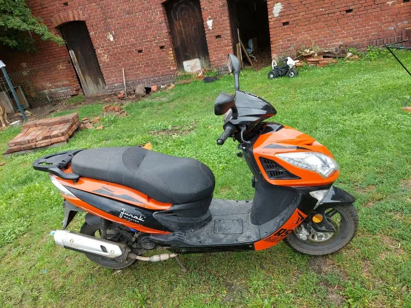 Pomarańczowo-czarny skuter marki Jurak zaparkowany na trawniku przed budynkiem z czerwonej cegły. Skuter jest ustawiony bokiem do obiektywu, posiada czarne siedzenie i srebrny wydech. W tle widoczne są drzwi w ceglanej ścianie oraz mały biały pojazd dziecięcy.