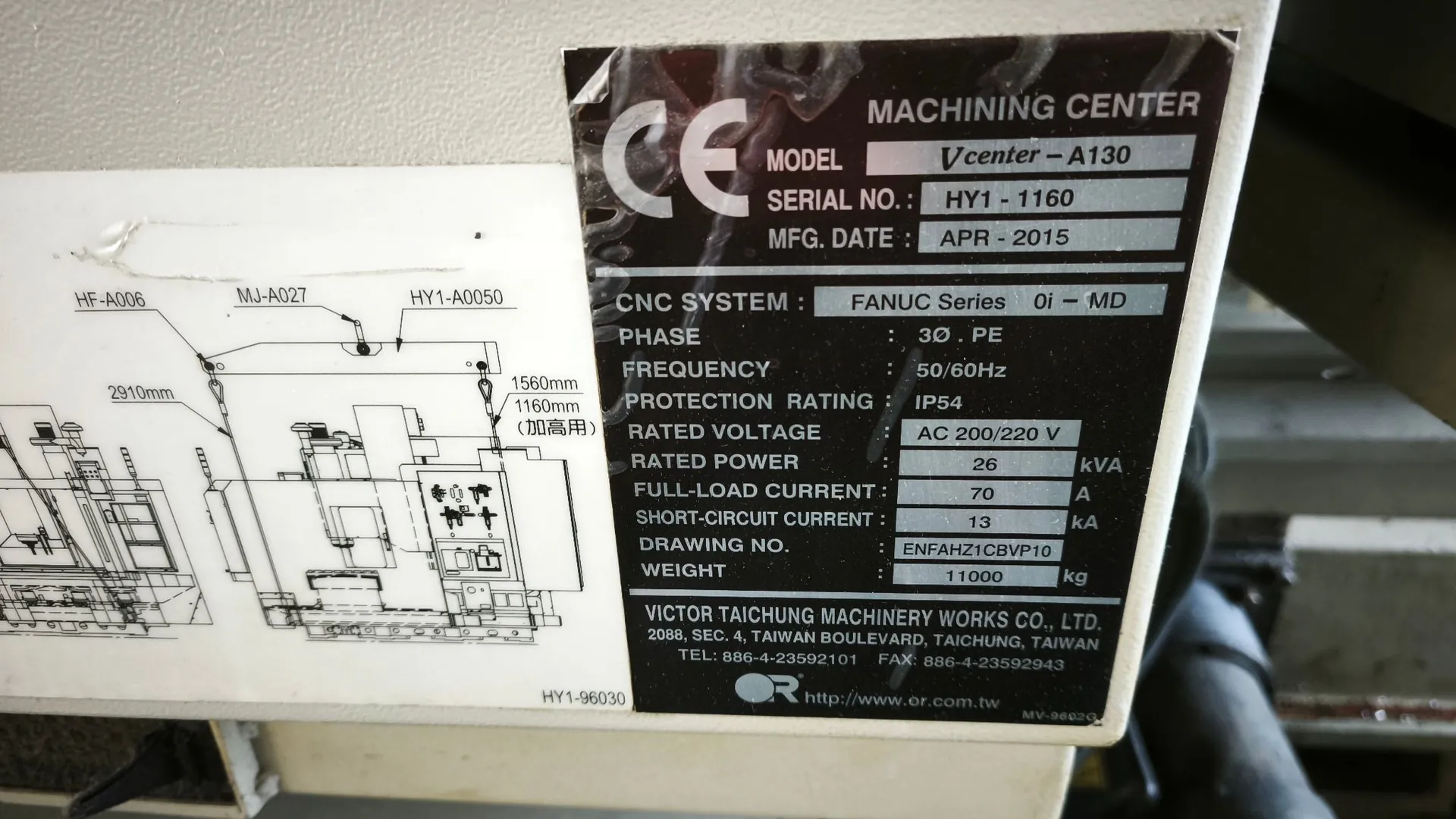Zdjęcie tabliczki znamionowej maszyny CNC. Na czarnym tle z białym tekstem widnieją następujące informacje: 'MACHINING CENTER', 'MODEL: Vcenter-A130', 'SERIAL NO.: HY1-1160', 'MFG. DATE: APR-2015'. Poniżej znajduje się sekcja techniczna: 'CNC SYSTEM: FANUC Series 0i-MD', 'PHASE: 3Φ PE', 'FREQUENCY: 50/60Hz', 'PROTECTION RATING: IP54', 'RATED VOLTAGE: AC 200/220 V', 'RATED POWER: 26 kVA', 'FULL-LOAD CURRENT: 70 A', 'SHORT-CIRCUIT CURRENT: 13 kA', 'DRAWING NO.: ENFAHZ1CBVP10', 'WEIGHT: 11000 kg'. Na dole widnieje nazwa producenta: 'VICTOR TAICHUNG MACHINERY WORKS CO., LTD.' wraz z adresem w Tajwanie, numerami telefonu i faksy oraz adresem strony internetowej http://www.or.com.tw. Po lewej stronie tabliczki znajduje się schemat techniczny maszyny z wymiarami, takimi jak 2910mm, 1560mm i 1160mm. W lewym górnym rogu tabliczki widoczne jest oznaczenie CE.