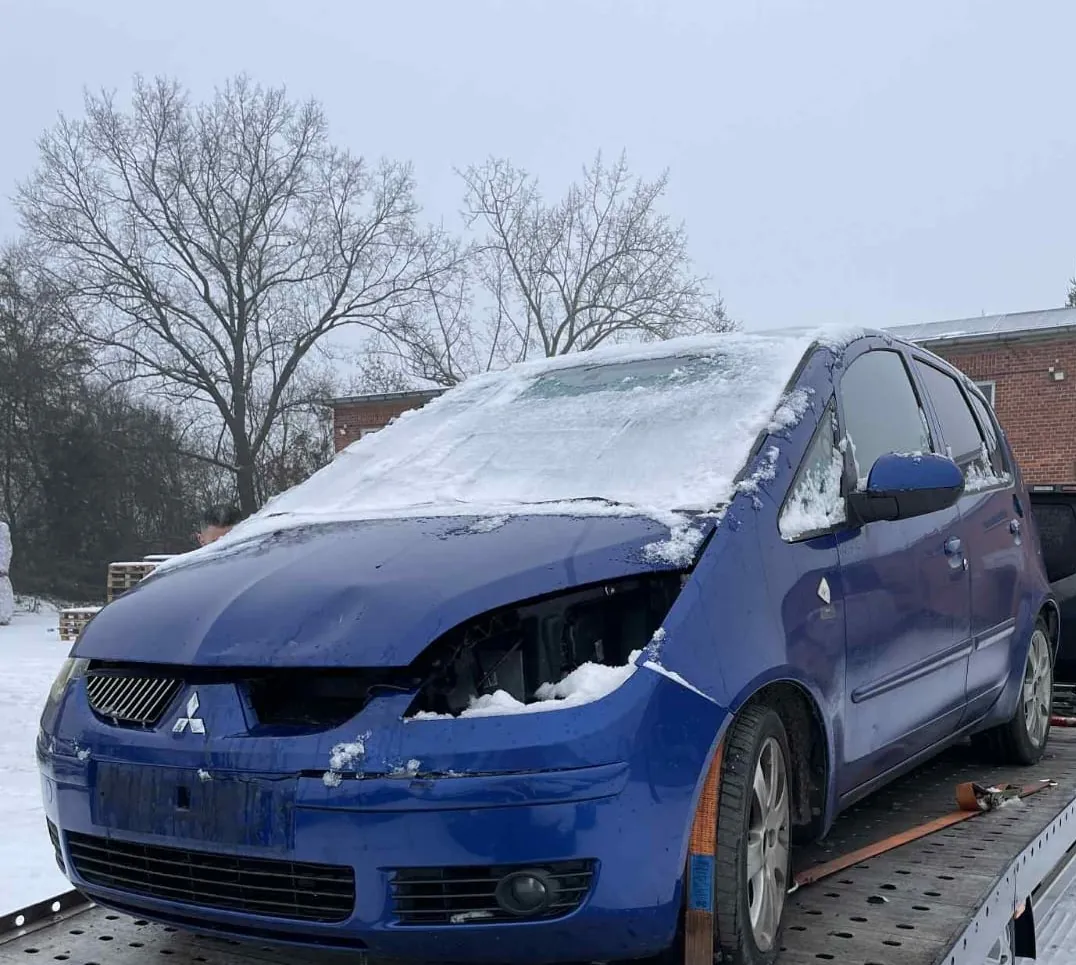 Niebieski samochód marki Mitsubishi z uszkodzonym przodem, pokryty śniegiem na masce i przedniej szybie. Samochód znajduje się na platformie holownika. W tle widoczne są bezlistne drzewa, ceglany budynek oraz śnieżna powierzchnia. Na masce widoczny jest logo Mitsubishi w postaci trzech rombów.
