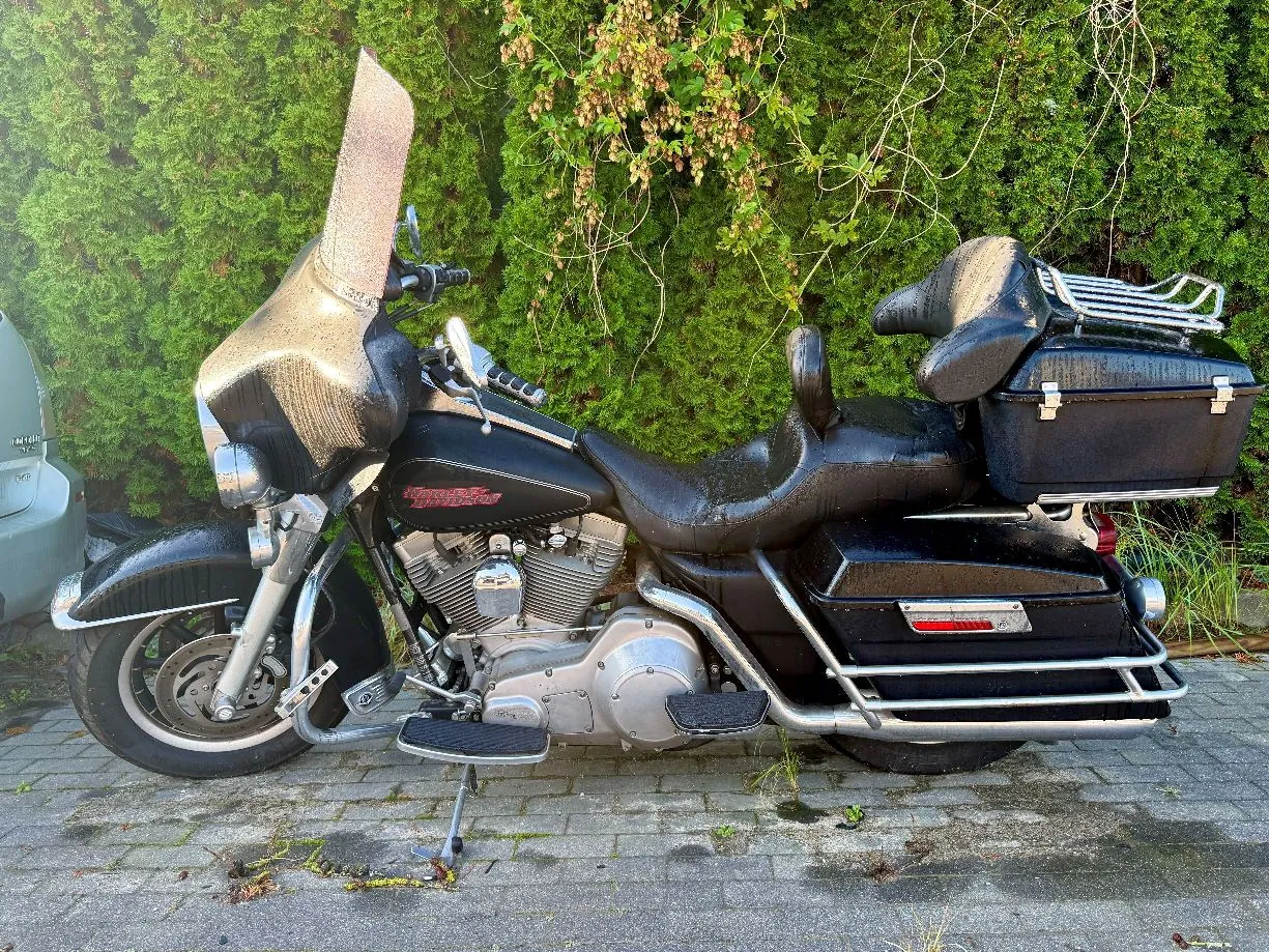Czarny motocykl typu touring z dużym przednimy ekranem, siedzeniem dla kierowcy i pasażera oraz dwiema skrzyniami bocznymi i jedną tylną. Pojazd stoi na kostce brukowej na tle zielonego żywopłotu. Motocykl jest mokry, z widocznymi kroplami wody na lakierze i siedzeniu.