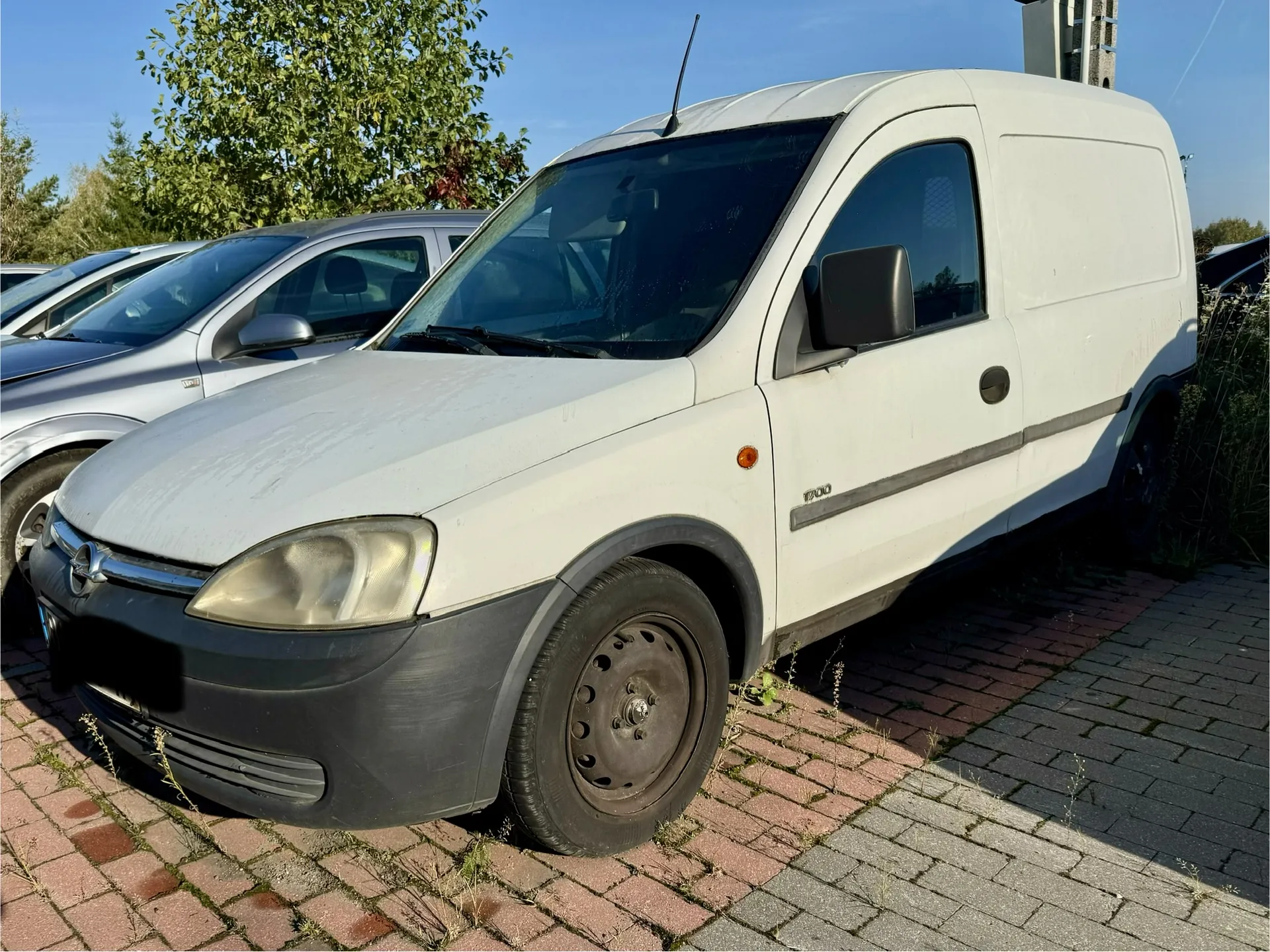 Biały samochód dostawczy marki Opel Combo zaparkowany na kostce brukowej. Pojazd jest widoczny z przodu i z boku. Obok niego stoi srebrny samochód osobowy. W tle widać zielone drzewa i fragment budynku pod jasnym, niebieskim niebem.