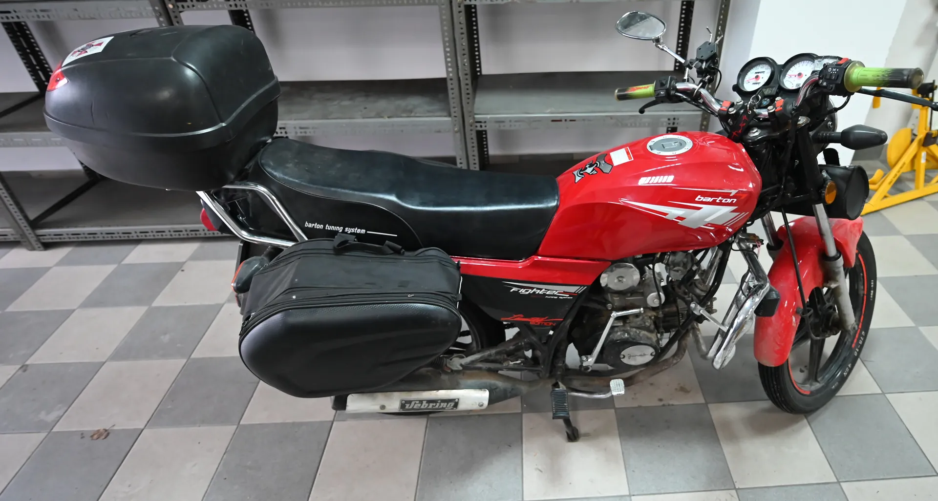 Czerwony motocykl marki Barton z czarnym siedzeniem, czarnym bagażnikiem i boczną torbą. Widoczne napisy: "Barton", "Barton tuning system", "Debrining". W tle metalowe półki i podłoga w szachownicę. Liczniki prędkości i zielone uchwyty na kierownicy.