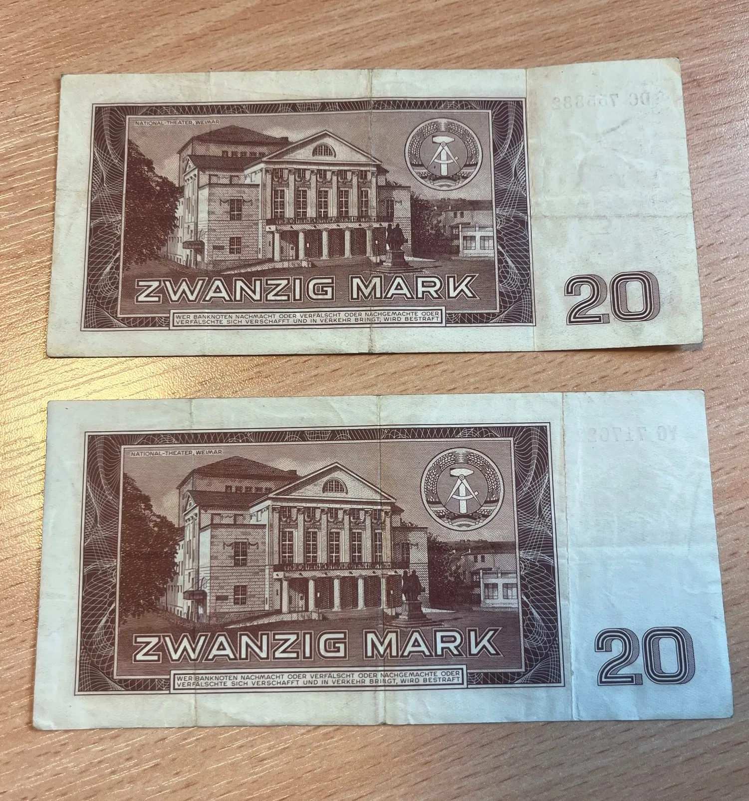 Dwa banknoty o nominale 20 marek (Zwanzig Mark). Na każdym z nich widnieje grafika przedstawiająca budynek National-Theater w Weimarze oraz symbol z młotem i cyrklem w okręgu. Na dole znajduje się napis 'ZWANZIG MARK' oraz mniejszy tekst ostrzegawczy dotyczący fałszerstw. Po prawej stronie widoczna jest duża cyfra '20'. Banknoty są ułożone jeden nad drugim na drewnianym blacie.