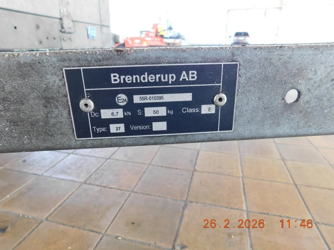 Zbliżenie na metalową tabliczkę z napisem "Brenderup AB" przykręconą do metalowej powierzchni dwoma śrubami. Na tabliczce widoczne są informacje: "E24", "55R-010395", "Dc: 6,7 kN", "S: 50 kg", "Class: E", "Type: 27", "Version: " (puste pole). W tle rozmyte elementy: betonowa konstrukcja, czerwony pojazd, samochód oraz podłoga z brązowymi płytkami. W prawym dolnym rogu pomarańczowy znaczek z datą i czasem: "26.2.2026 11:46".