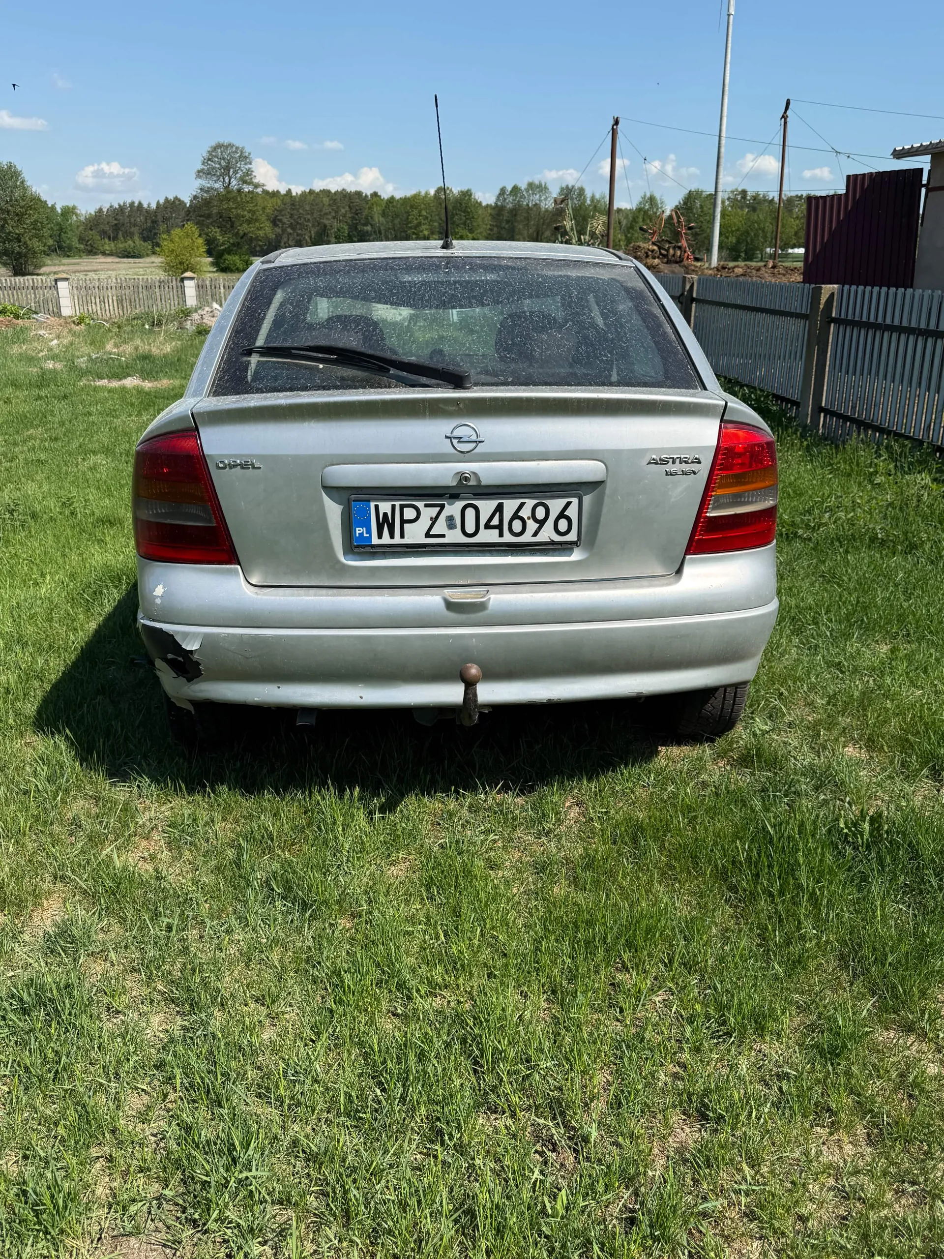 Srebrny samochód Opel Astra widziany od tyłu, z tablicą rejestracyjną WPZ 04696 (PL). Widoczne uszkodzenie lewego tylnego zderzaka oraz hak holowniczy. Samochód stoi na trawie, w tle drewniane ogrodzenie, drzewa, słupy energetyczne i budynek. Niebo jest jasne z kilkoma chmurami.