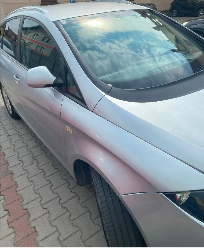 Srebrny samochód osobowy zaparkowany na kostce brukowej. Zdjęcie przedstawia przednią prawą część pojazdu, w tym maskę, przednie prawe koło, lusterko boczne oraz przednią szybę, w której odbija się niebo z chmurami.