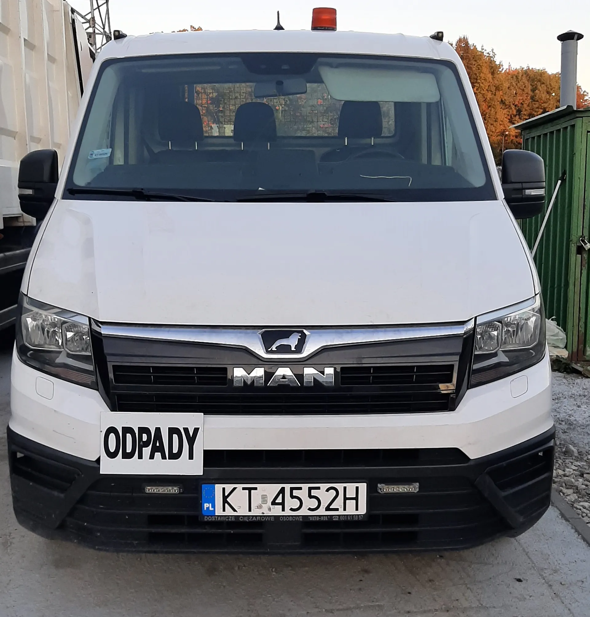 Biały samochód dostawczy marki MAN z przodu. Na masce widoczny napis "MAN" oraz logo lwa. Poniżej tablica rejestracyjna z polskim kodem "PL" i numerem "KT 4552H". Nad tablicą biały znak z czarnym napisem "ODPADY". Na dachu czerwone światło ostrzegawcze. W tle zielony kontener, drzewa z jesiennymi liśćmi oraz fragment innego pojazdu po lewej stronie. Samochód stoi na betonowej powierzchni.