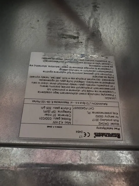 Zdjęcie tabliczki znamionowej pieca katalitycznego marki Ravanson. Tabliczka zawiera następujące informacje: Model: OKG42, Rok produkcji: 2017, Nr seryjny: 00052, Kraj przeznaczenia: PL. Specyfikacja techniczna: Moc: 4,2 kW, Rodzaj gazu: G30/G31, Ciśnienie: 37 mbar, Kategoria: I3(P) (B/P), Zużycie gazu: 305 g/h. Widnieje napis: RAVANSON LTD Sp z o.o. - ul. Mazowiecka 6, 09-100 Płońsk oraz numer normy: N. P. EN 443. Poniżej znajduje się sekcja 'UWAGA' z instrukcjami bezpieczeństwa dotyczącymi wentylacji i użytkowania urządzenia.