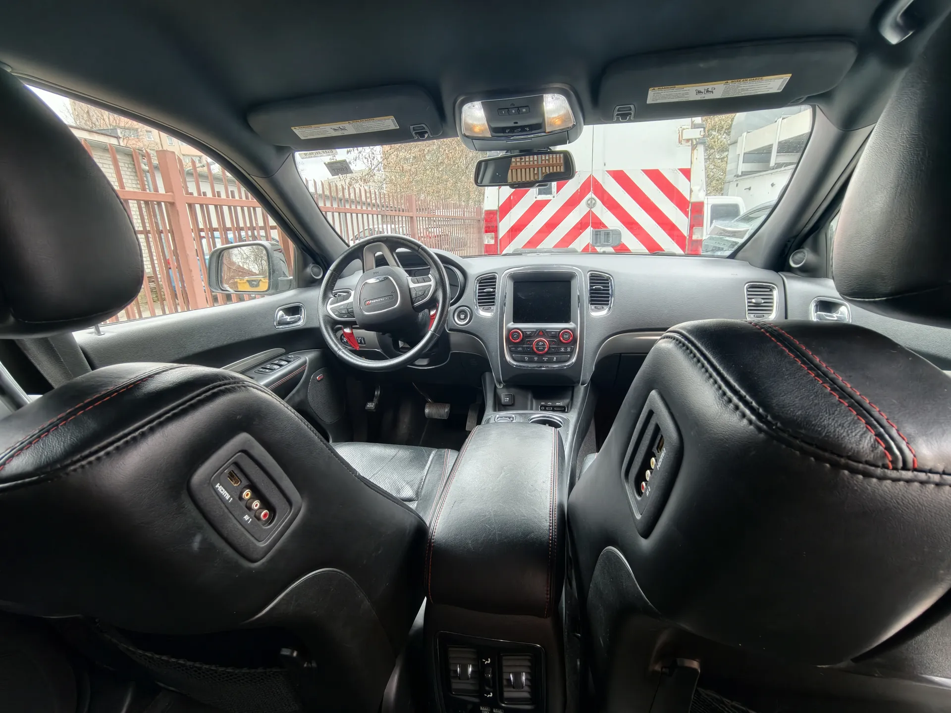 Wnętrze samochodu widziane z tyłu przednich siedzeń. Czarne skórzane fotele z czerwonym przeszyciem, kierownica z logo, centralna konsoła z ekranem i czerwonymi przyciskami, lusterko wsteczne oraz daszki przeciwsłoneczne z widocznymi etykietami. Przez przednią szybę widać czerwono-białą barierę, ogrodzenie i budynki na zewnątrz.