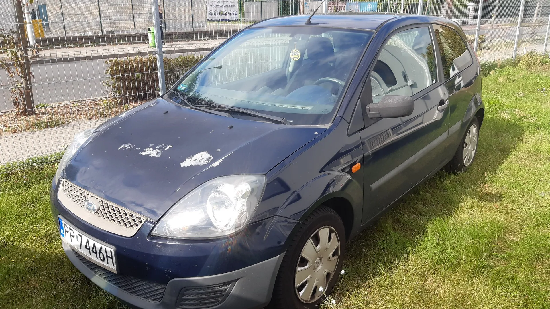 Granatowy samochód osobowy marki Ford zaparkowany na trawie obok metalowego ogrodzenia. Pojazd ma widoczne uszkodzenia lakieru na masce. Tablica rejestracyjna samochodu to PP 7446H.