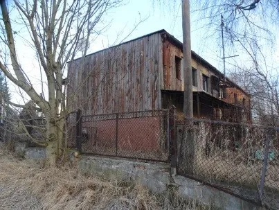 Stary, dwukondygnacyjny budynek z drewnianą elewacją w kolorze brązowym i szarym. Budynek ma kilka małych okien i znajduje się za metalowym ogrodzeniem z siatki osadzonej na betonowym murku. Wokół budynku rosną bezlistne drzewa i sucha trawa.