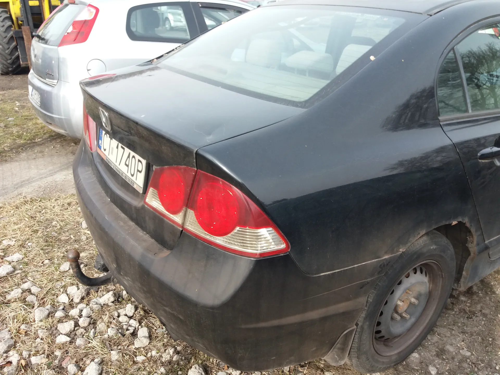 Zdjęcie przedstawia tył czarnego samochodu marki Honda z tablicą rejestracyjną CT 1740P. Samochód stoi na podłożu z kamieniami i suchą trawą. W tle widoczny jest srebrny samochód oraz fragment żółtego pojazdu. Czarny samochód ma zabrudzoną karoserię, czerwone światła tylne i brak koła na tylnej osi. Tablica rejestracyjna jest czytelna i umieszczona poziomo.