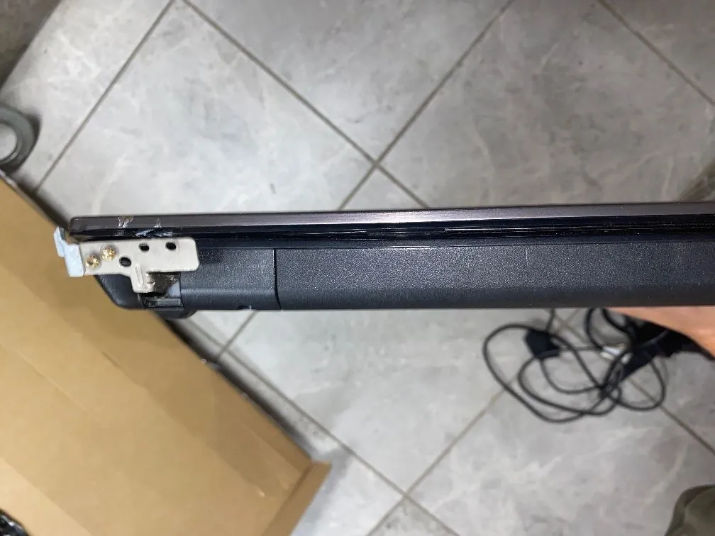 Zbliżenie na krawędź czarnej obudowy laptopa, w której widoczne jest uszkodzenie mechaniczne. Fragment plastikowej ramki jest wyrwany, odsłaniając metalowy zawias i śruby mocujące. Urządzenie znajduje się na tle szarej płytki ceramicznej, obok brązowego kartonu.