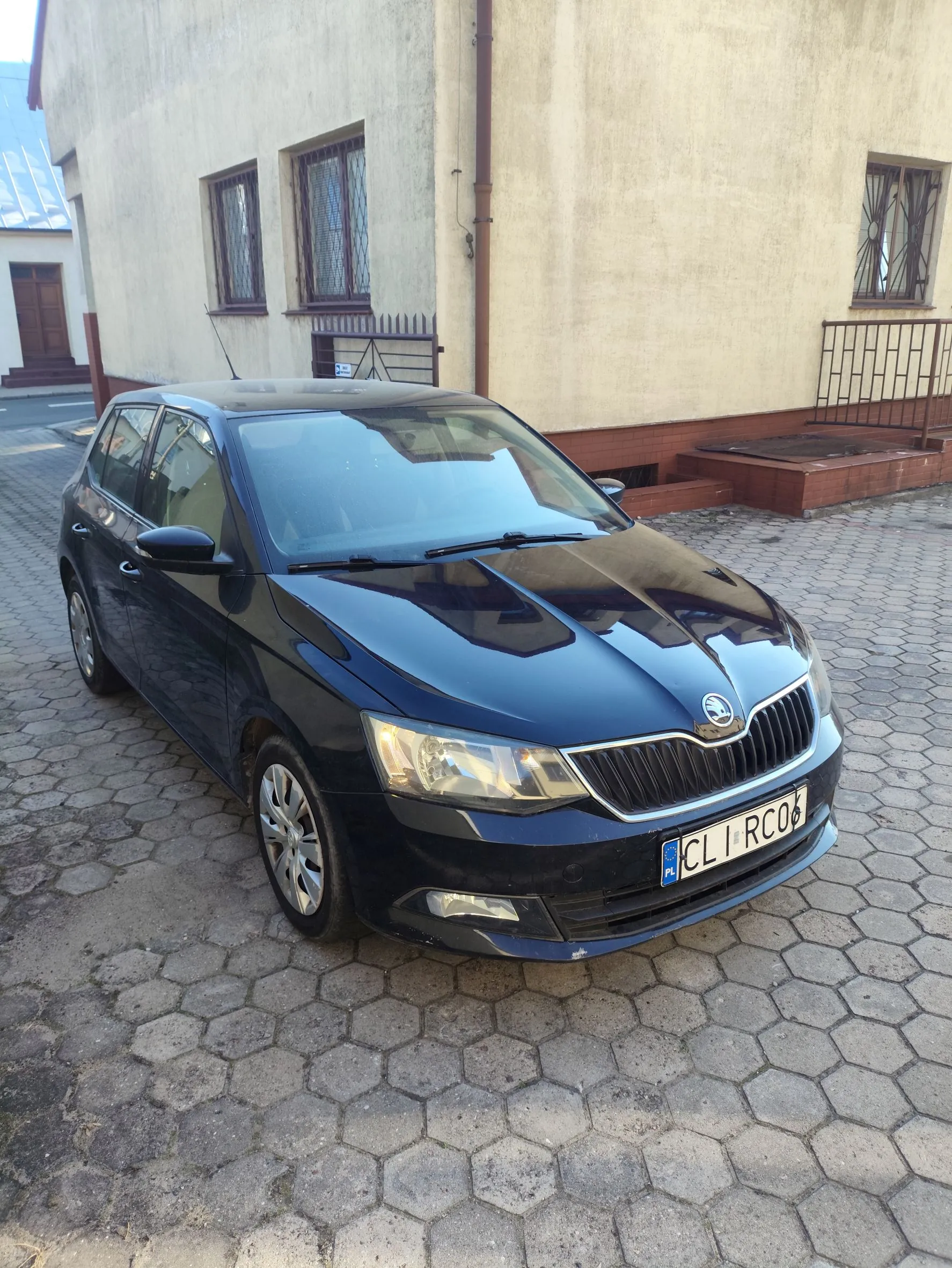 Ciemnoniebieski samochód marki Skoda zaparkowany na podjeździe z szarej, sześciokątnej kostki brukowej. Auto stoi przed jasnym budynkiem z oknami zabezpieczonymi kratami. Na samochodzie widoczna jest polska tablica rejestracyjna o numerze CLI RC06.