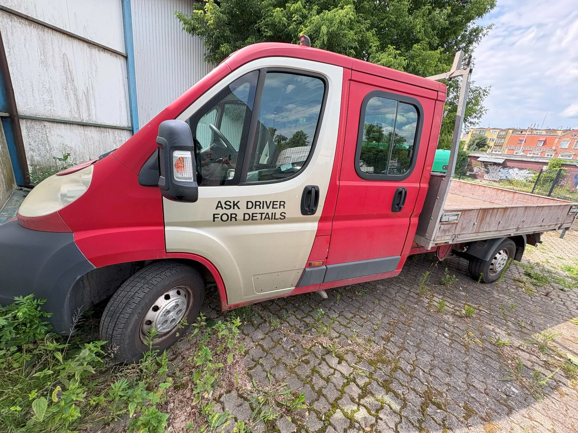 Czerwony samochód dostawczy typu chassis z otwartą platformą ładunkową. Pojazd ma beżowe drzwi kierowcy i pasażera, na których znajduje się czarny napis: 'ASK DRIVER FOR DETAILS'. Samochód stoi zaparkowany na kostce brukowej porośniętej trawą, w tle widoczne są zielone drzewa oraz fragmenty budynków.