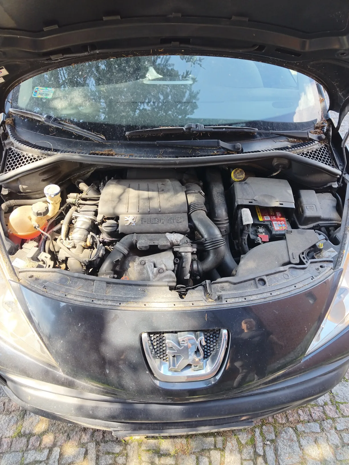 Widok z góry na otwartą maskę czarnego samochodu marki Peugeot, ukazującą silnik spalinowy oraz elementy komory silnika, takie jak akumulator i zbiornik z płynem chłodniczym. Na przodzie maski widoczne jest chromowane logo lwa Peugeot.