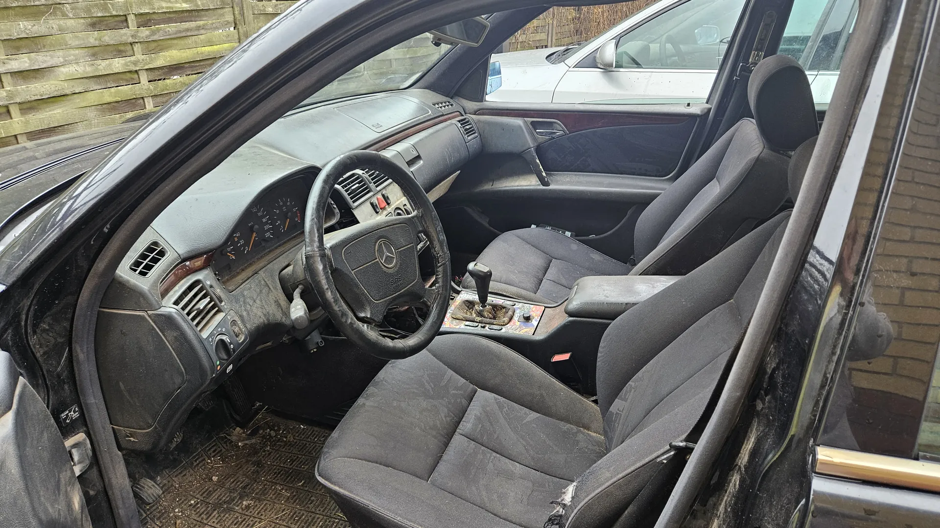 Wnętrze samochodu z otwartą drzwią pasażera. Widoczna czarna kierownica z logo Mercedes-Benz, zniszczona tapicerka siedzeń, deska rozdzielcza z licznikami i przyciskami, oraz centralna konsoła z przyciemnionym pokrętłem biegów. W tle drewniane ogrodzenie, inny samochód oraz ceglana ściana. Całość pokryta warstwą kurzu i brudu.