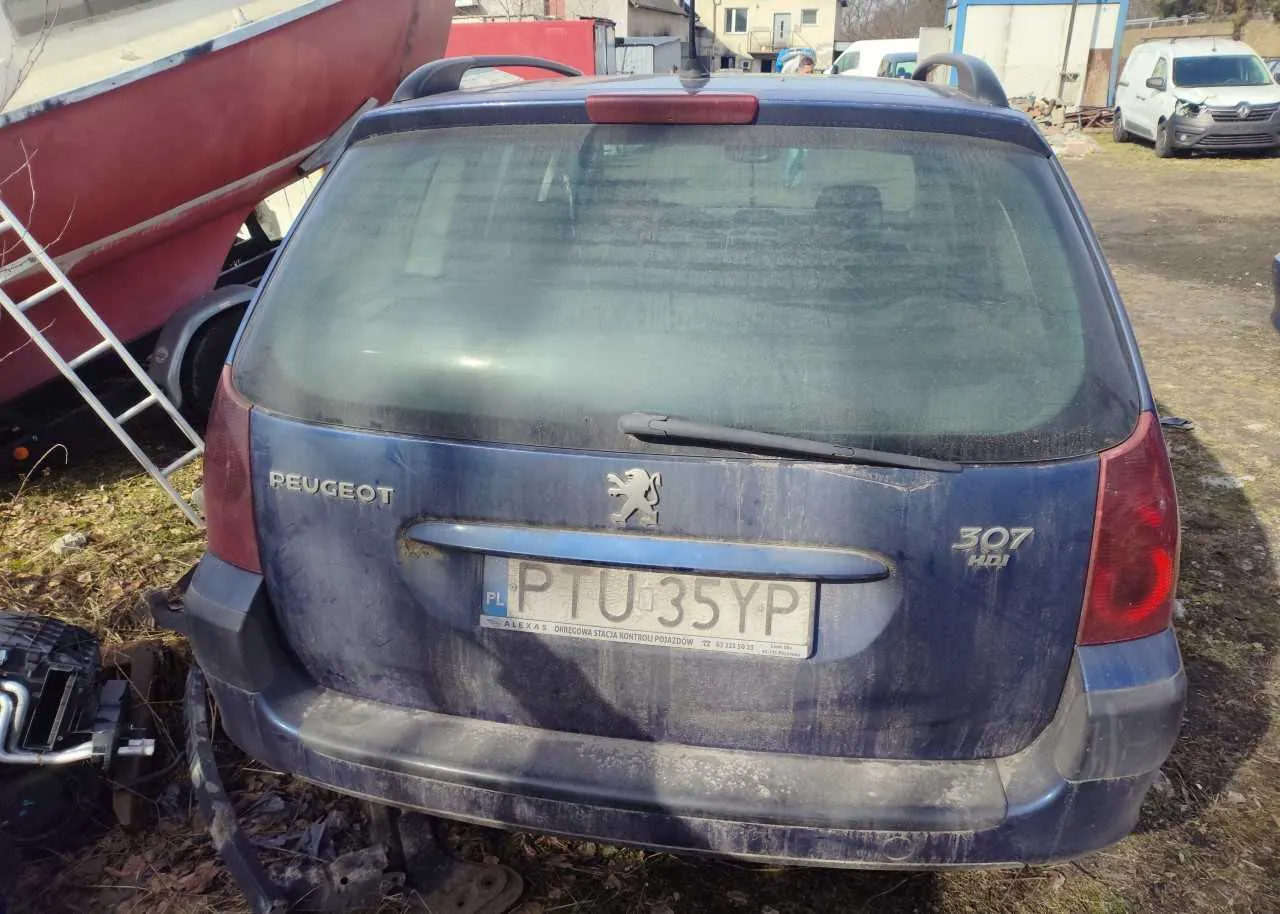 Zdjęcie przedstawia tył niebieskiego samochodu Peugeot 307 HDI z zabrudzoną karoserią. Na bagażniku widoczne są napisy 'PEUGEOT', logo lwa oraz '307 HDI'. Tablica rejestracyjna PL PTU 35YP z napisem 'ALEXAS OŚRODEK SZKOLENIA KIEROWCÓW' na dole. Po lewej stronie samochodu znajduje się czerwona łódź na przyczepie z metalową drabiną opartą o nią. W tle widoczne są inne pojazdy, w tym biały furgon, oraz budynki. Podłoga pokryta suchą trawą i liśćmi.