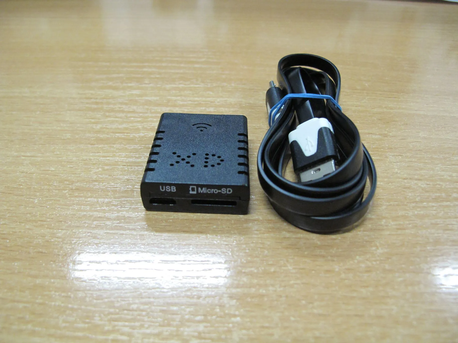 Czarny, niewielki adapter sieciowy z napisem 'USB' oraz 'Micro-SD' na przednim panelu i ikoną sygnału Wi-Fi na górze. Obok urządzenia znajduje się zwinięty czarny kabel USB z niebieską opaską zabezpieczającą. Przedmioty leżą na jasnobrązowej, drewnianej powierzchni.