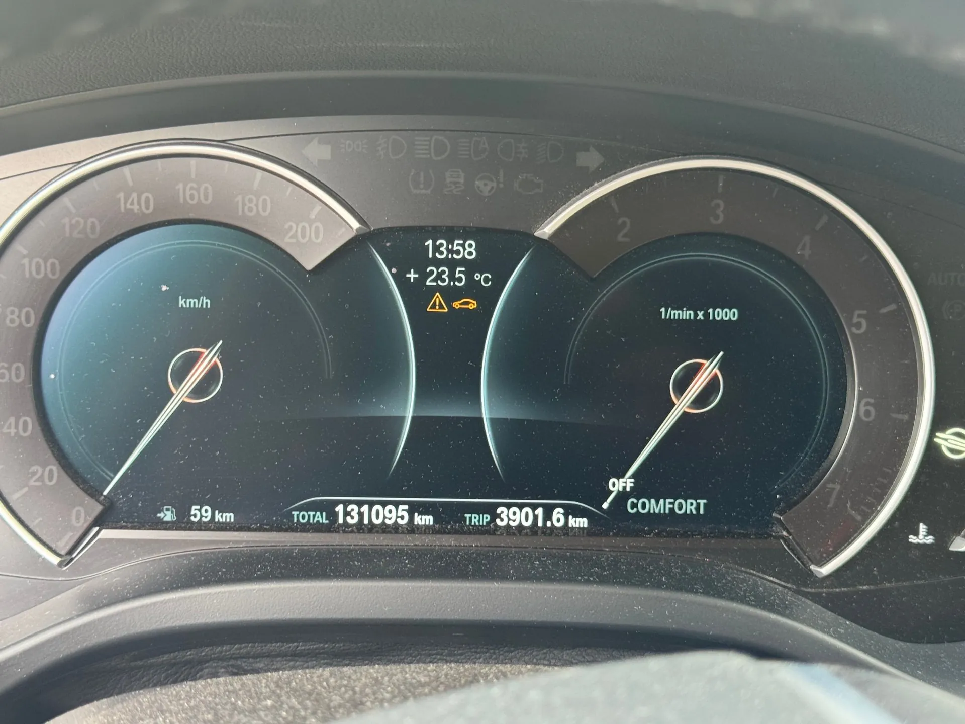 Zdjęcie deski rozdzielczej samochodu z cyfrowymi wskaźnikami. Po lewej stronie znajduje się prędkościomierz z zaznaczoną prędkością 0 km/h, po prawej obrotomierz z napisem '1/min x 1000'. W centralnej części wyświetlacza widnieje godzina 13:58, temperatura +23.5 °C oraz ikona ostrzegawcza z samochodem. Na dole ekranu widoczne są dane: zasięg 59 km, całkowity przebieg TOTAL 131095 km oraz przebieg podróży TRIP 3901.6 km. Po prawej stronie widnieje napis 'OFF COMFORT'.