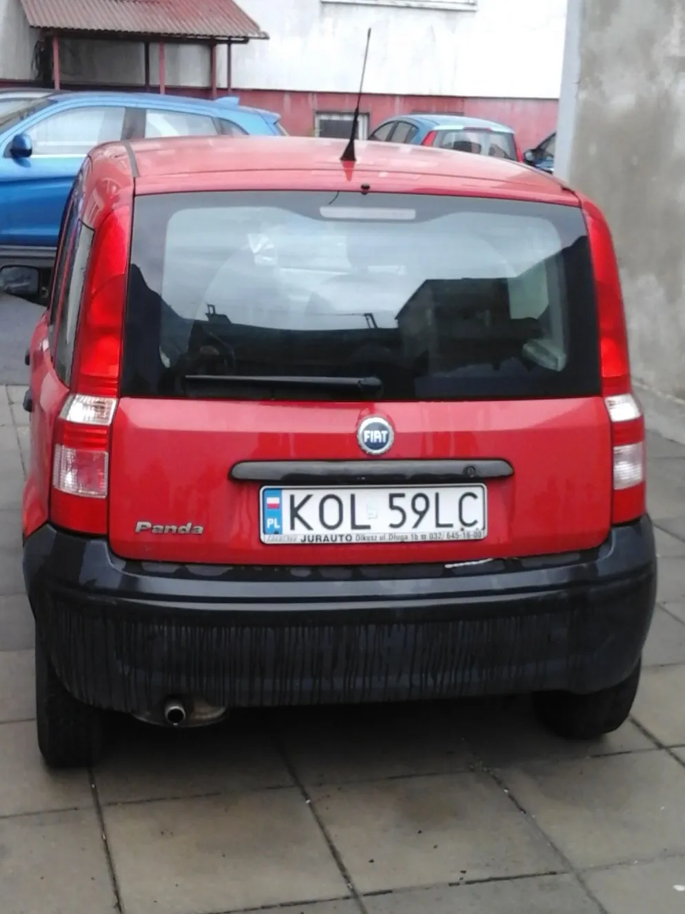 Tylny widok czerwonego samochodu marki Fiat Panda. Pojazd posiada czarny zderzak i polską tablicę rejestracyjną o numerze KOL 59LC. Poniżej tablicy widnieje napis 'JURAUTO Dikecet z Dluga 16 m 032/ 645-16-00'. W tle widoczne są inne zaparkowane samochody oraz fragmenty budynków.