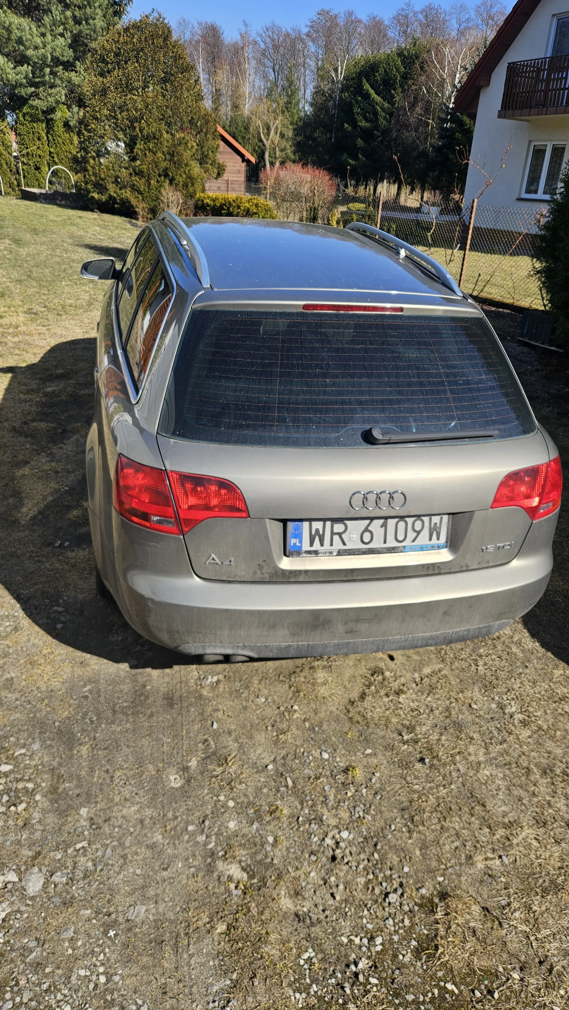 Srebrny samochód Audi A4 widziany od tyłu. Tablica rejestracyjna z numerem WR 6109W. Samochód zaparkowany na podwórku z suchą trawą i żwirem. W tle dom, drzewa i ogrodzenie. Niebo jest bezchmurne.
