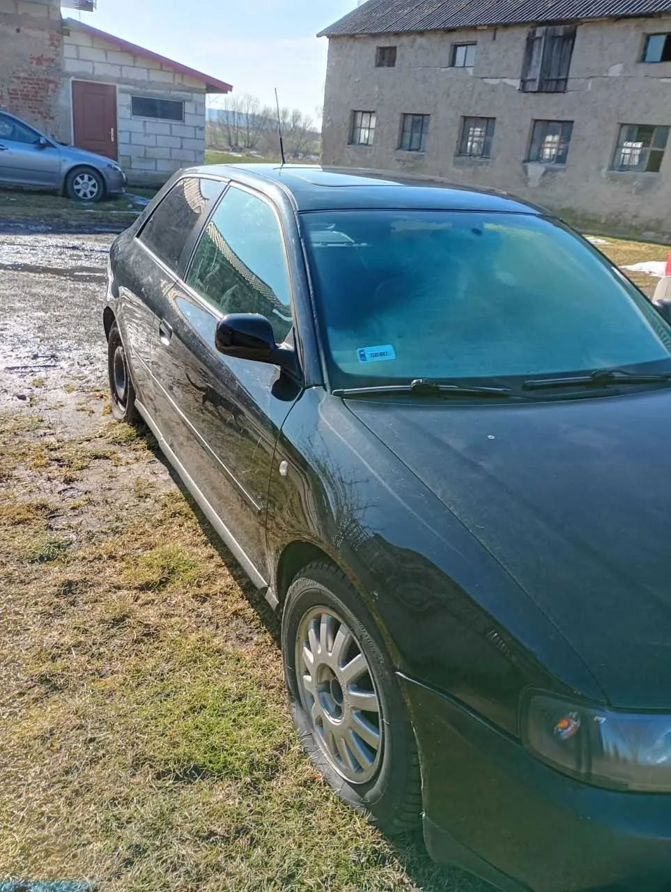 Ciemnozielony samochód osobowy typu hatchback zaparkowany na terenie z trawą i błotem. Na przedniej szybie widoczna niebieska naklejka. W tle dwa budynki mieszkalne: po lewej budynek z cegły z czerwonymi drzwiami i srebrnym samochodem, po prawej starszy, betonowy budynek z uszkodzonymi oknami. Na pierwszym planie widoczne koło z aluminiową felgą oraz lusterko boczne. Tło uzupełnia jasne niebo i suche drzewa w oddali.