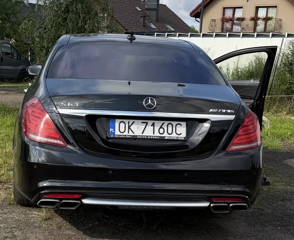 Tylny widok czarnego samochodu Mercedes-Benz klasy S AMG. Pojazd posiada polską tablicę rejestracyjną o numerze OK 7160C. Na klapie bagażnika widoczne są oznaczenia 'S 63' po lewej stronie oraz 'AMG' po prawej, a w centrum logo marki Mercedes. Samochód ma podwójne końcówki układu wydechowego po obu stronach z dolnym srebrnym dyfuzorem. Drzwi pasażera są otwarte. W tle widoczne są drzewa oraz fragmenty budynków mieszkalnych.