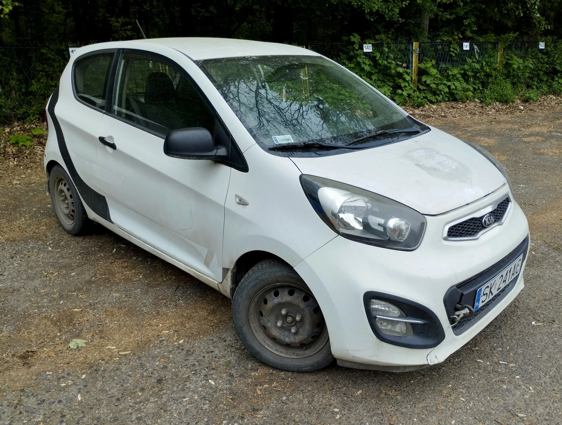 Biały samochód marki Kia o nadwoziu hatchback, zaparkowany na szutrowym podłożu na tle zielonych drzew. Pojazd ma czarne lusterka boczne i czarne elementy wykończenia w dolnej części drzwi. Na przednim zderzaku widoczna jest tablica rejestracyjna o numerze SK 241AG.